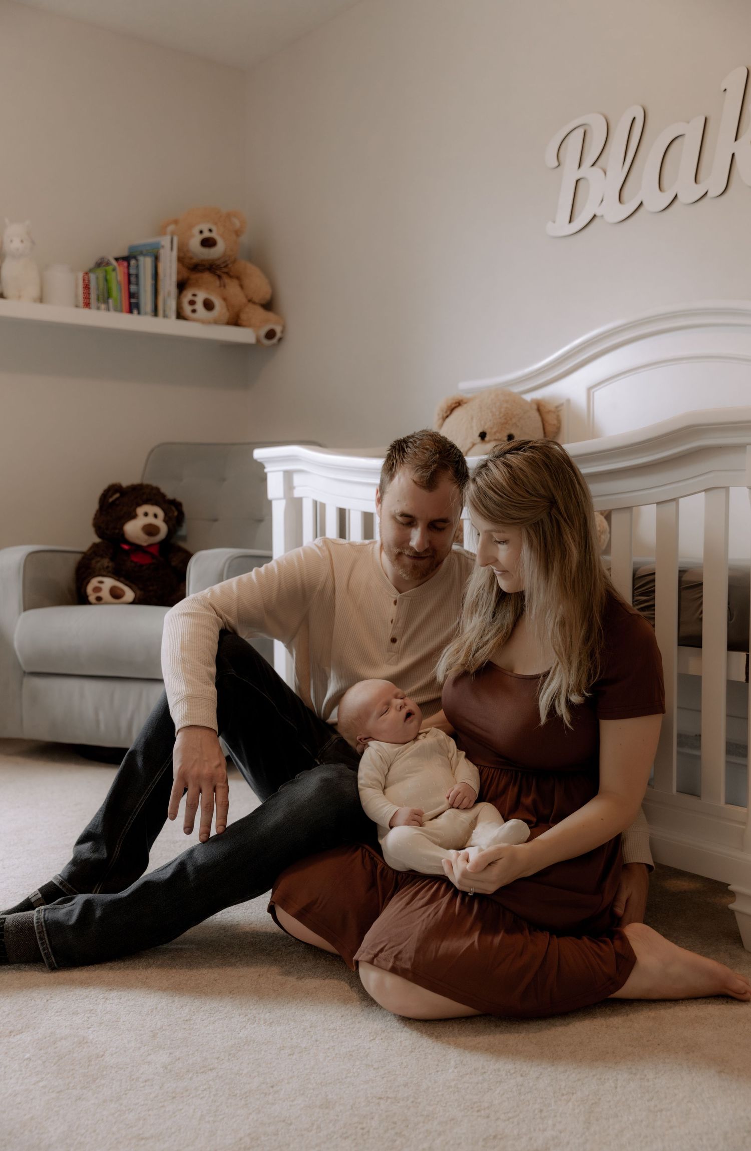 Intimate family moment in a nursery with stuffed animals on shelves as parents sit on floor with their newborn.