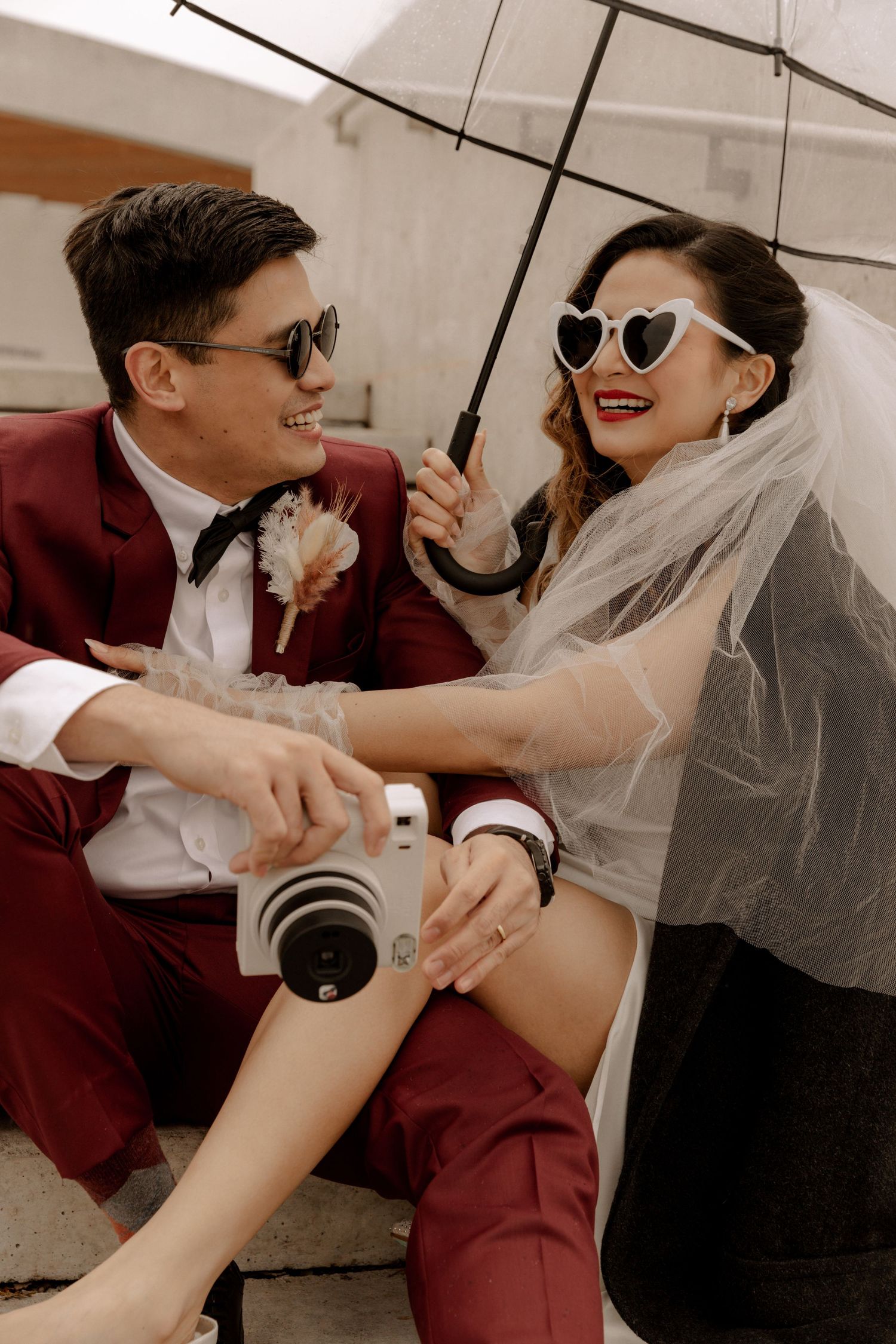 Playful wedding photoshoot with couple sharing umbrella and vintage camera.