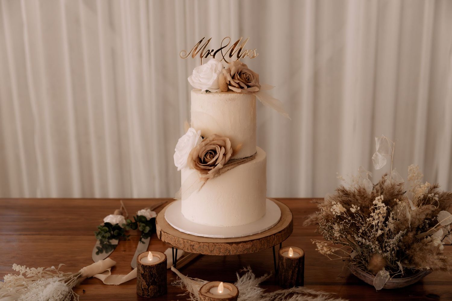 Elegant white wedding cake with gold accents and dried floral decorations on wooden stand.