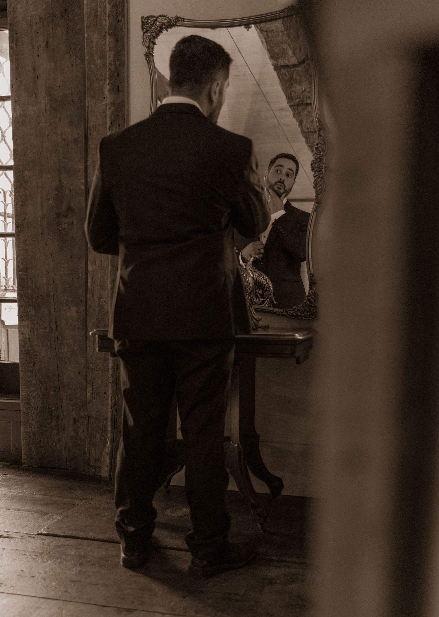 Sepia photograph of groom adjusting tie while looking in ornate mirror.