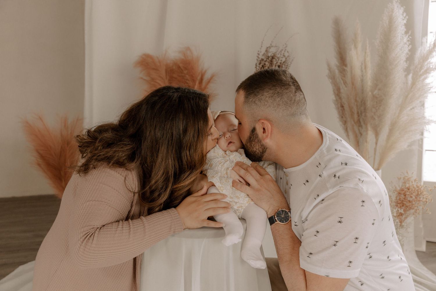 Couple leaning in together with baby between them, surrounded by pampas grass decor in a soft warm light.