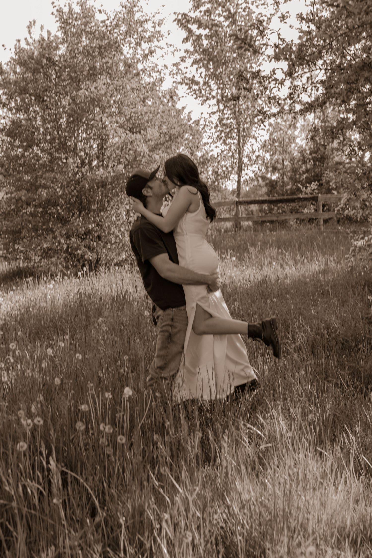 A romantic couple embracing in a grassy meadow with trees in the background, captured in sepia tones.
