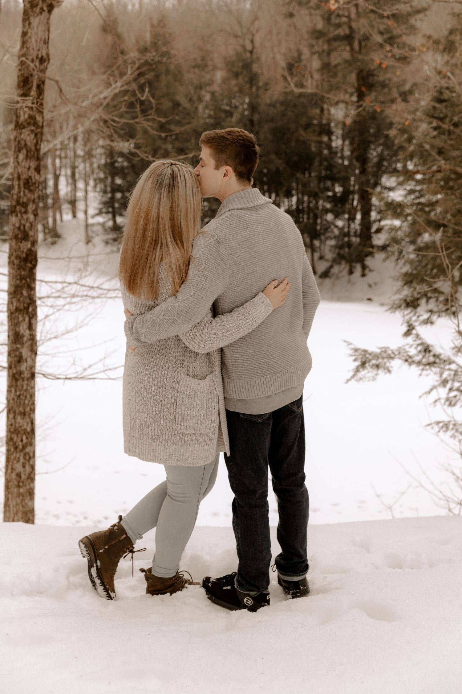 Two people share an intimate winter moment in cozy sweaters while standing in snow-covered woods.