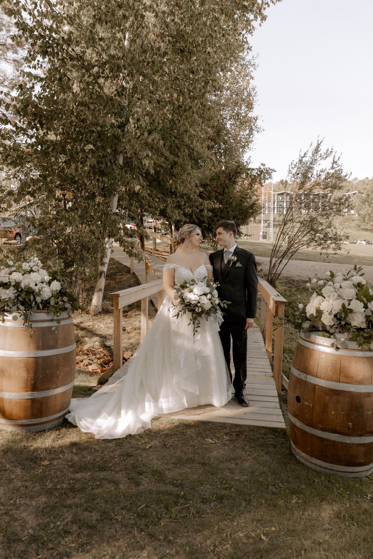 Wedding couple poses on wooden bridge decorated with wine barrels and floral arrangements.