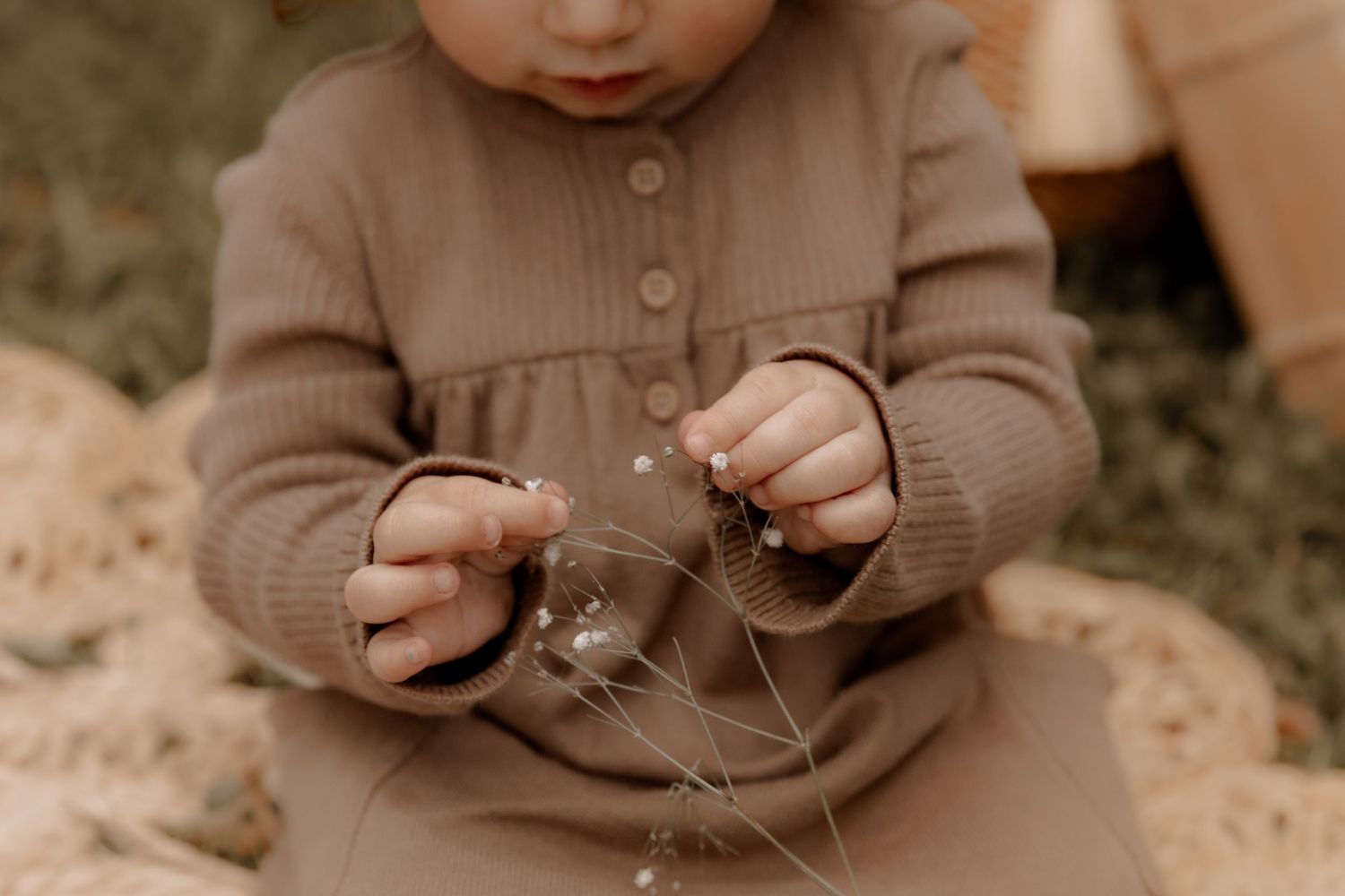 Small hands in a brown sweater dress carefully examining something in the grass.