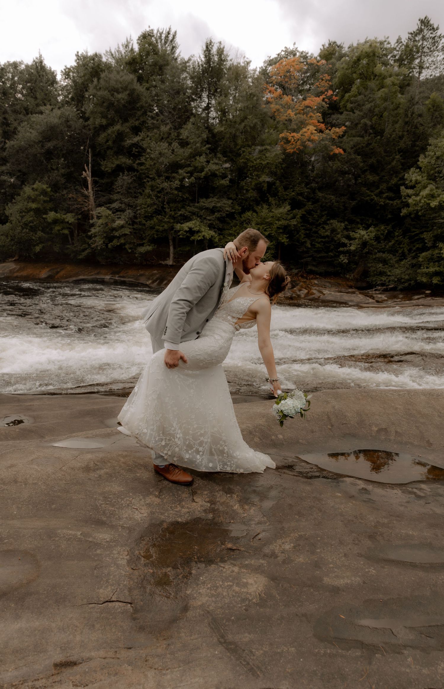 Newlyweds share romantic dip kiss near rushing waterfall in forest setting.