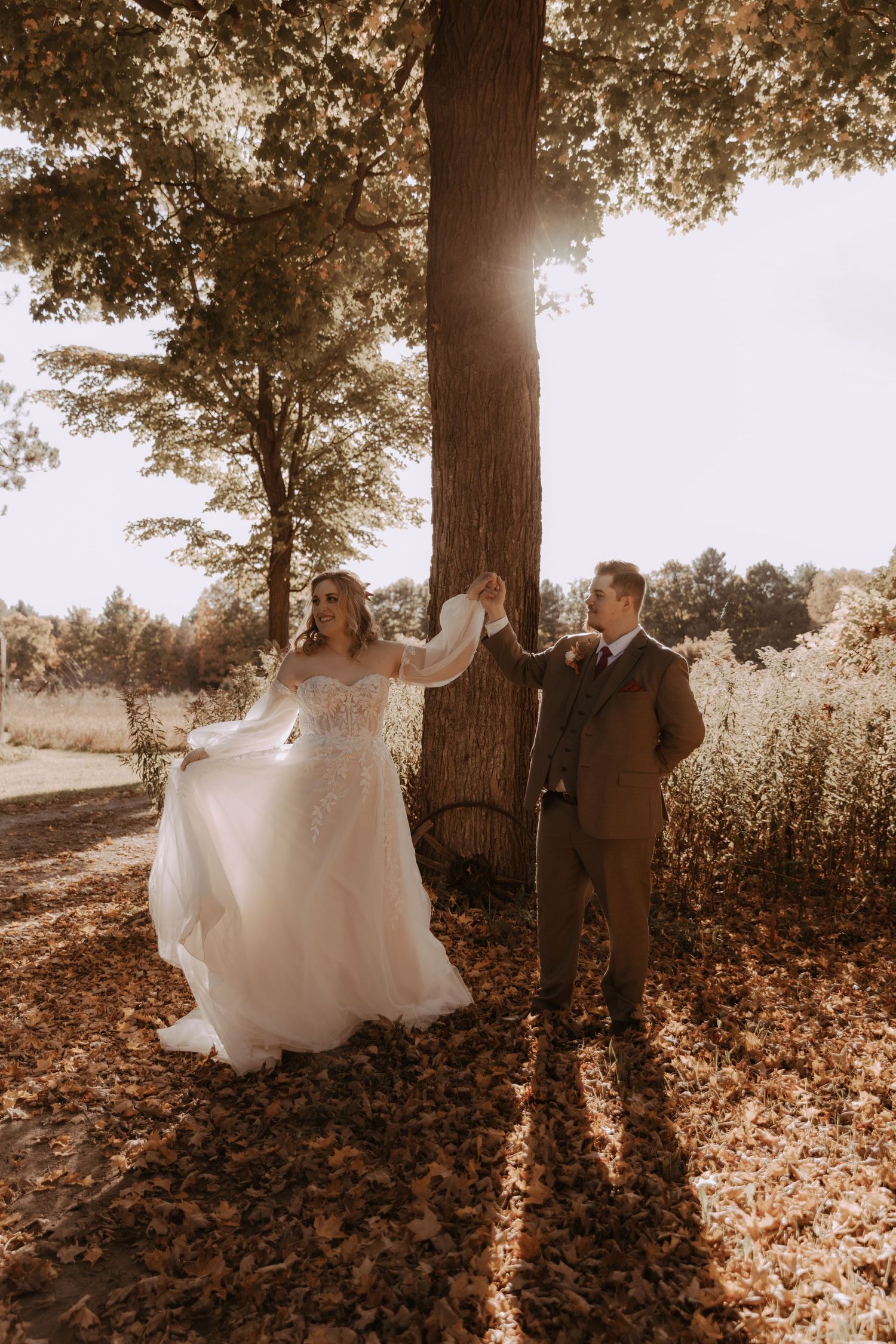 Bride in flowing gown and groom pose beneath a majestic tree with sunlight streaming through branches.