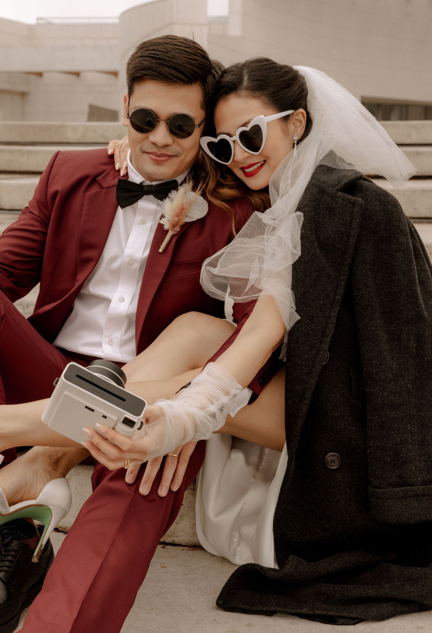 Modern wedding couple with vintage camera wearing heart sunglasses and burgundy suit.