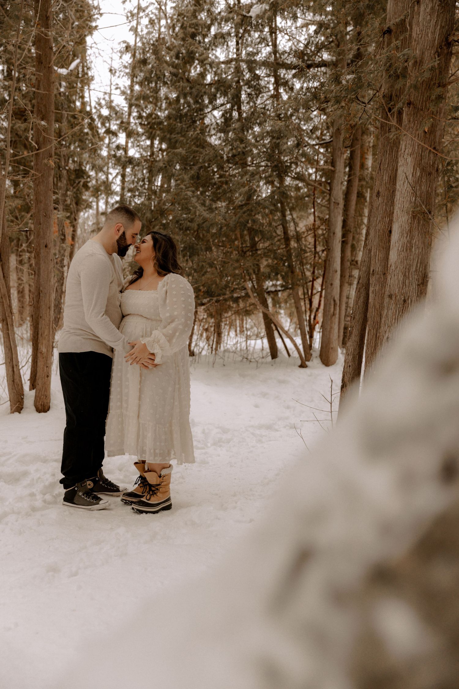 A couple in warm winter clothing shares an intimate moment on a snowy path through a pine forest.