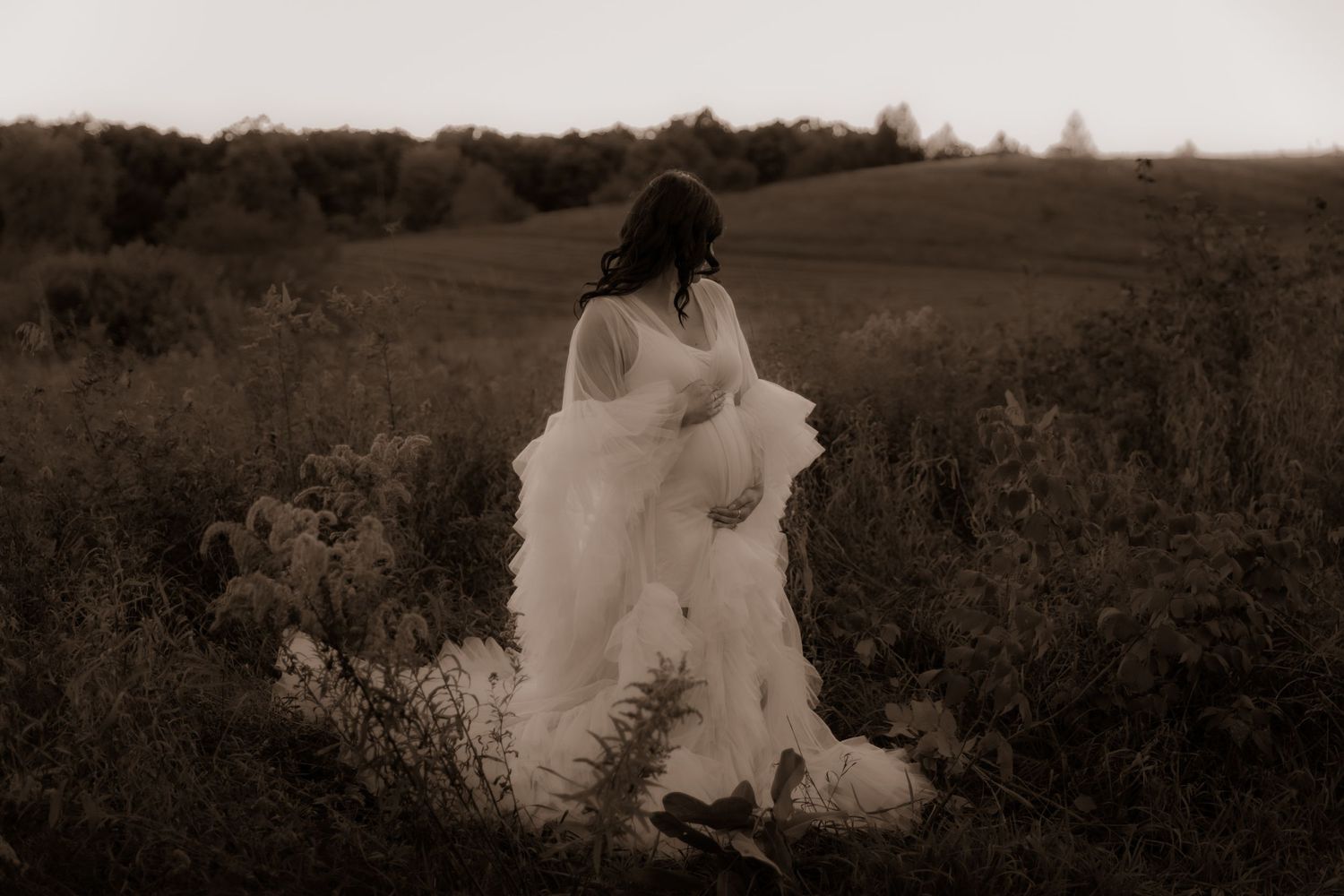 A sepia-toned figure in a flowing white dress stands among wild grasses in a rural field at dusk.