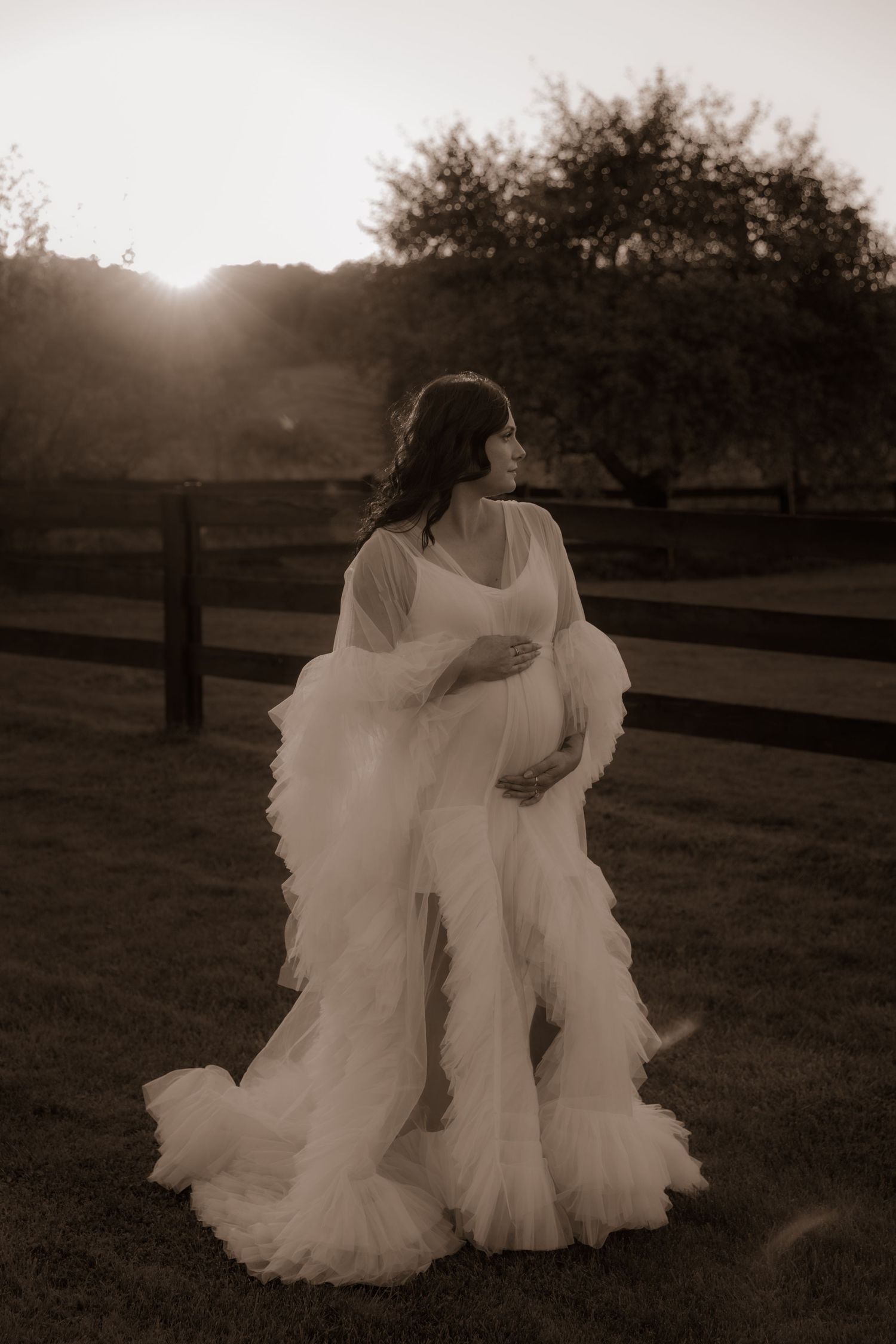 Ethereal sepia-toned maternity portrait featuring a billowing white dress against rustic farm fencing at golden hour.