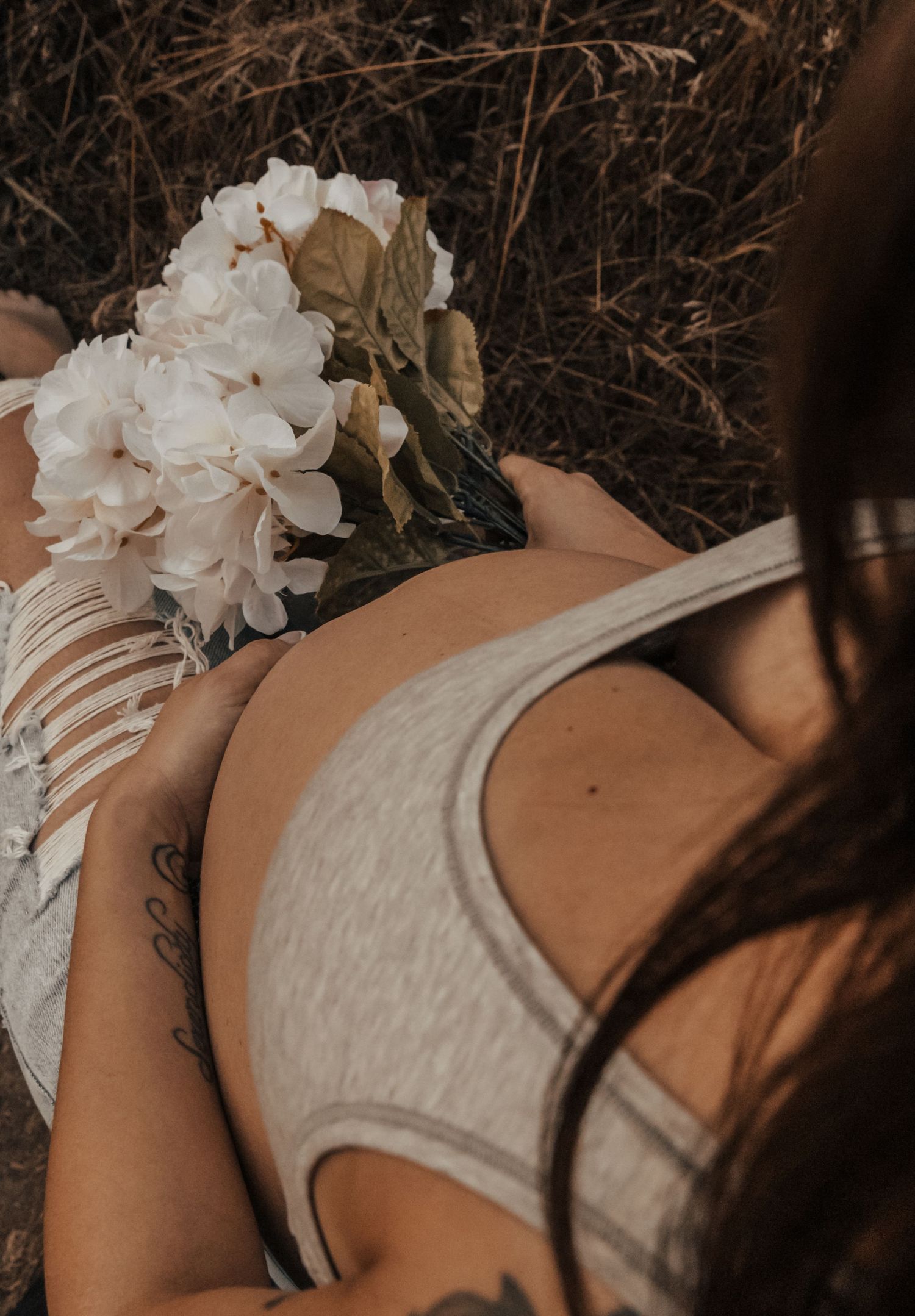 A person in a gray tank top holding white flowers poses outdoors against a dark natural background.