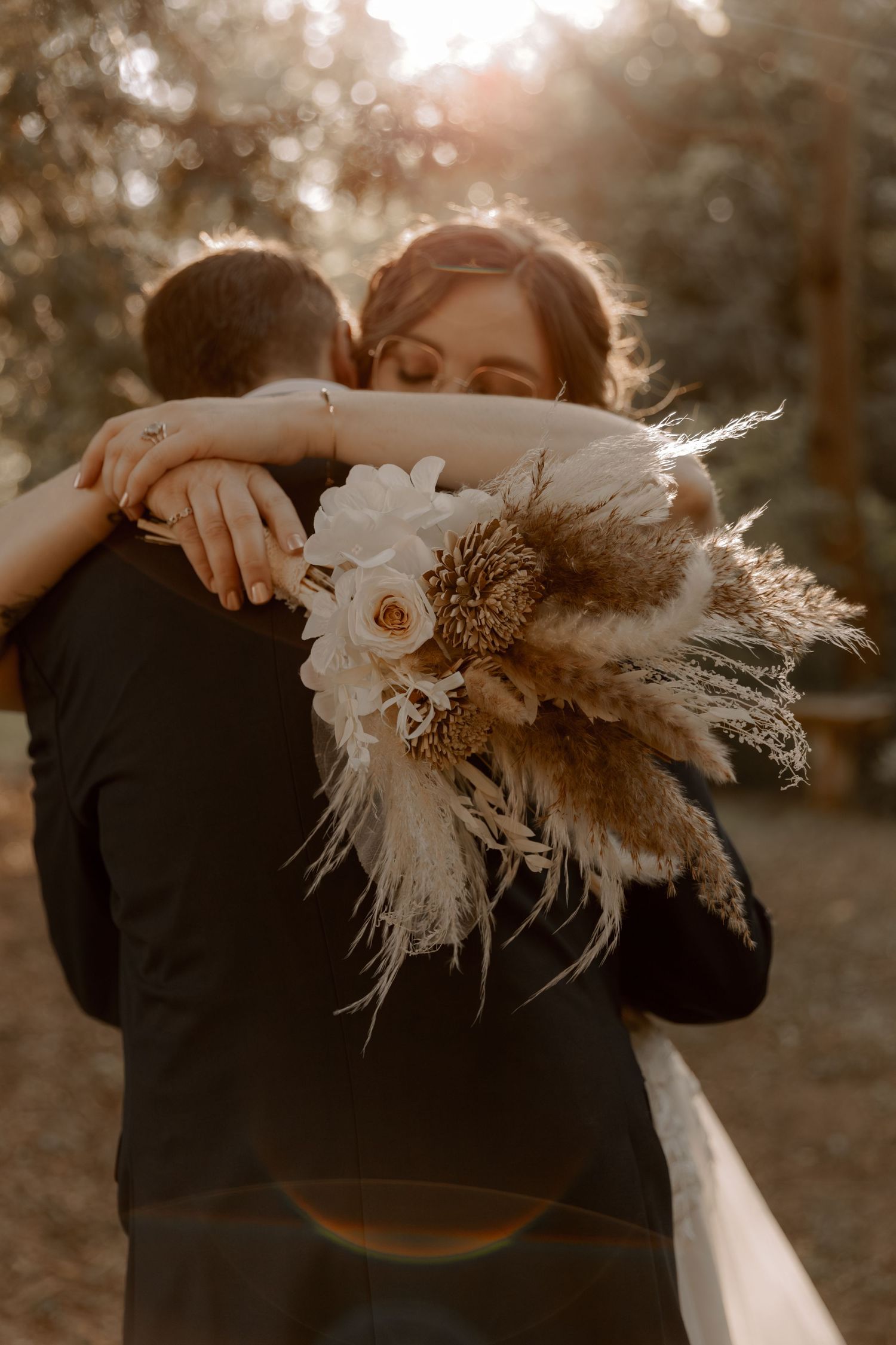 A couple embraces at sunset while holding a beautiful dried pampas grass and floral wedding bouquet.