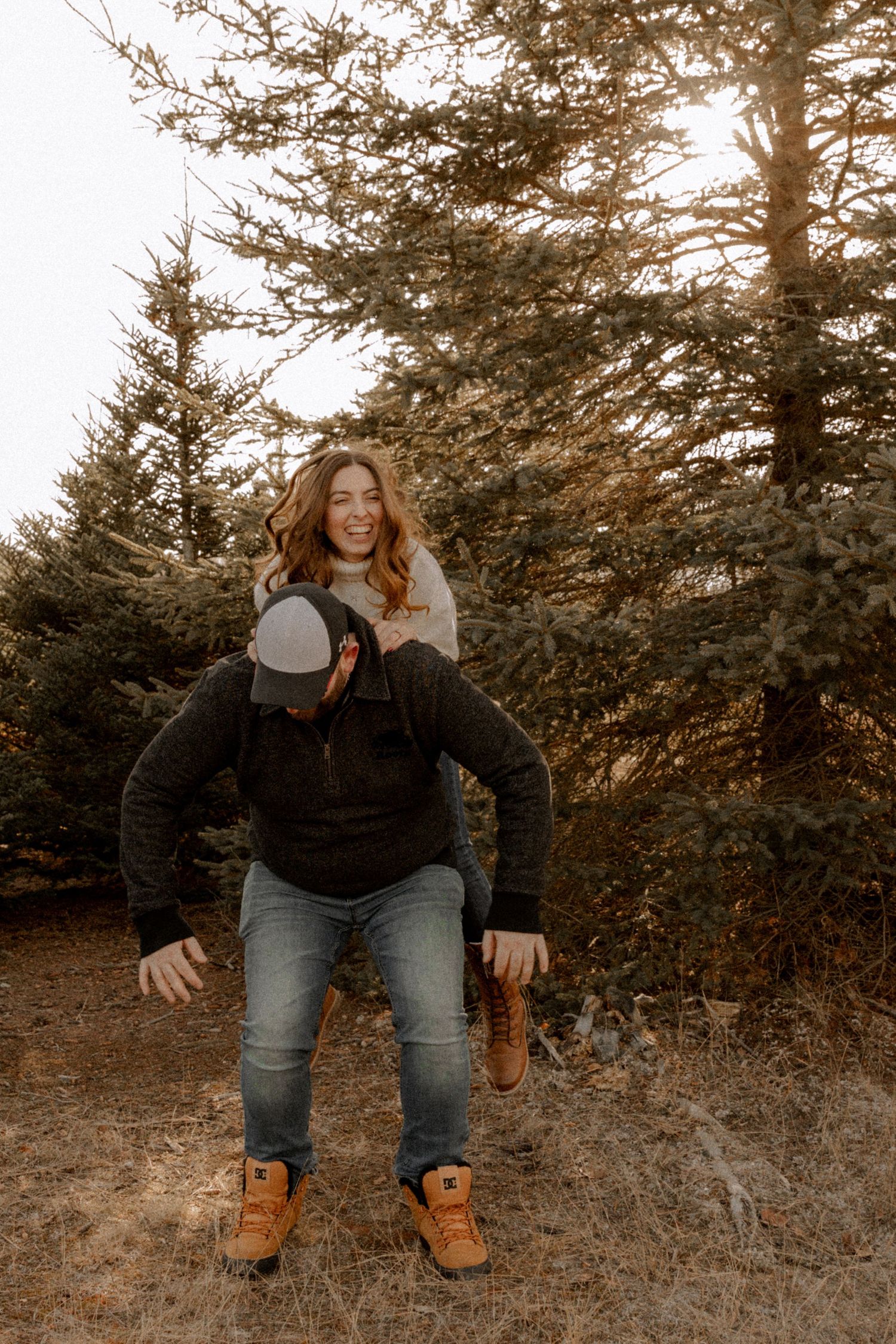 Person in a black fleece jacket and jeans poses outdoors among evergreen trees at sunset with warm lighting.