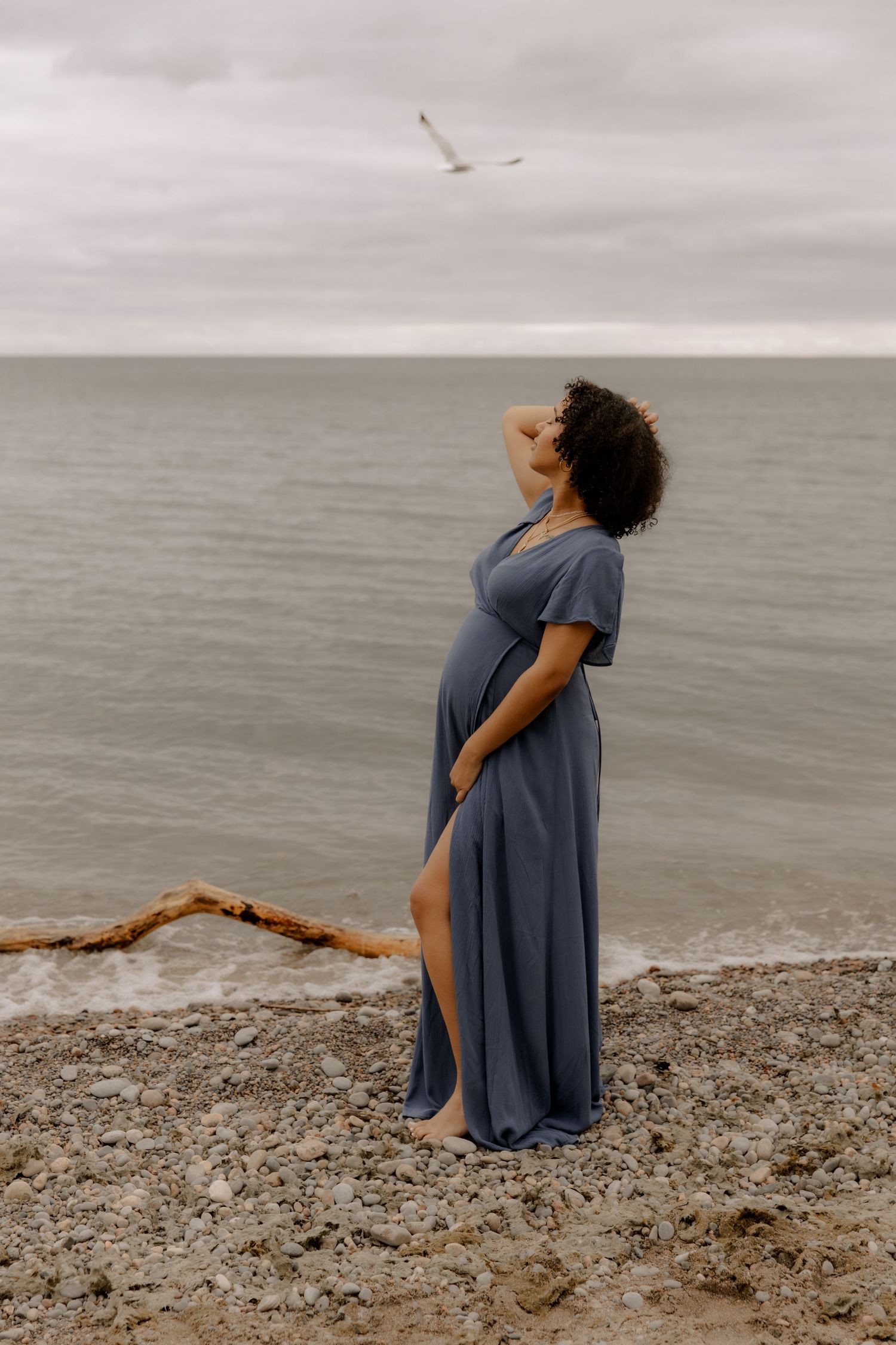 A pregnant woman in a blue dress stands on a beach watching a seagull fly overhead.