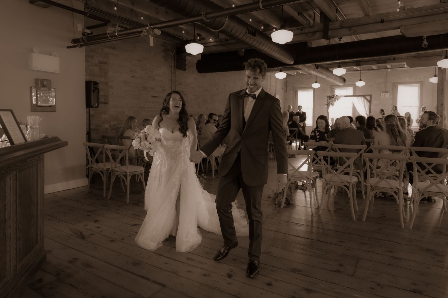 A joyful wedding processional photographed in sepia tones within a beautifully lit brick venue.
