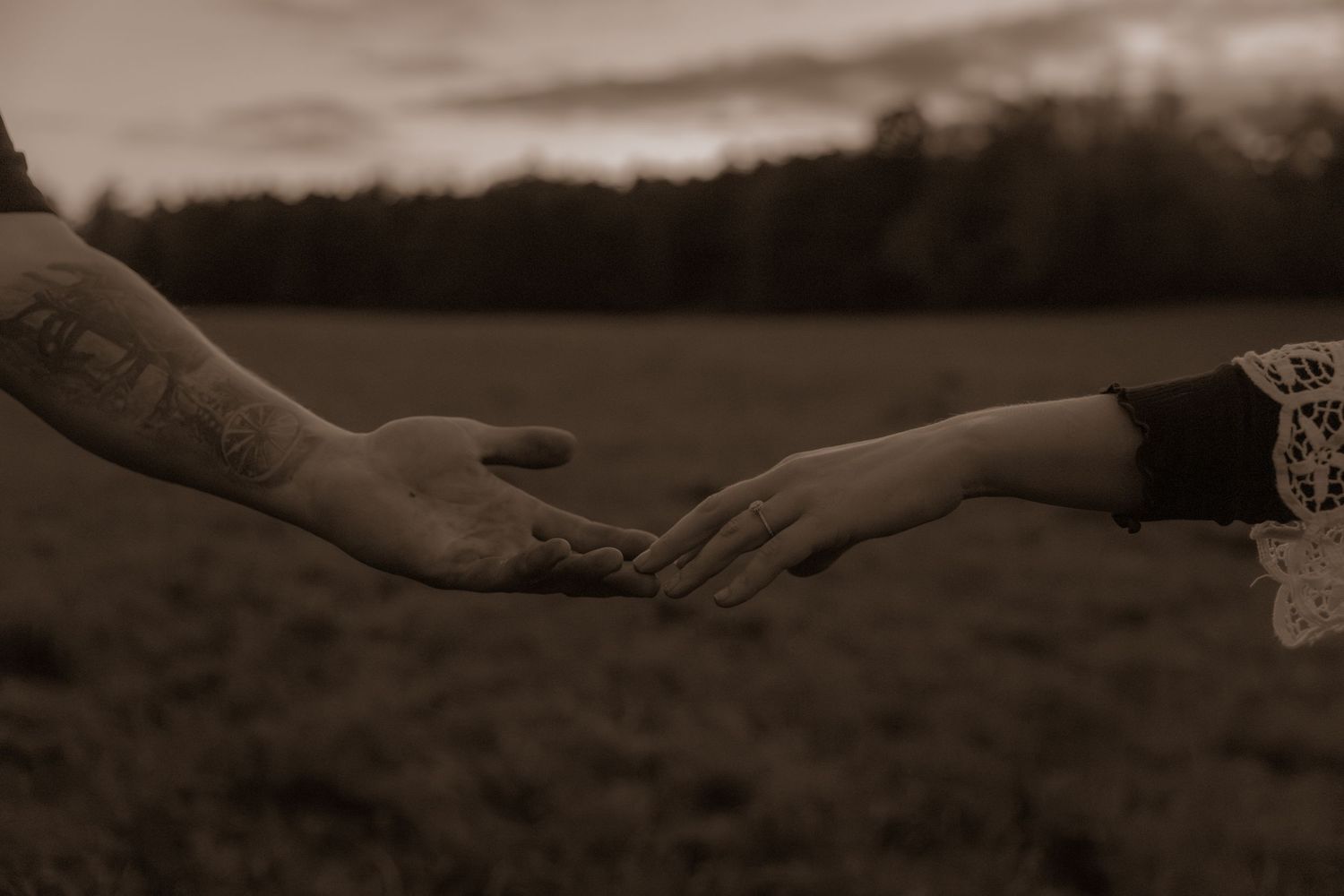 Close up of two hands gently touching in a romantic gesture against a moody outdoor backdrop.