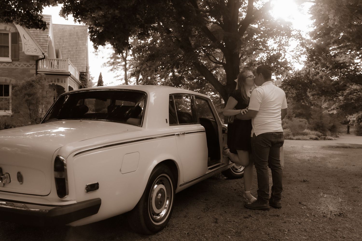A vintage Rolls Royce or luxury car parked in a driveway with someone standing beside the passenger door in sepia tones.
