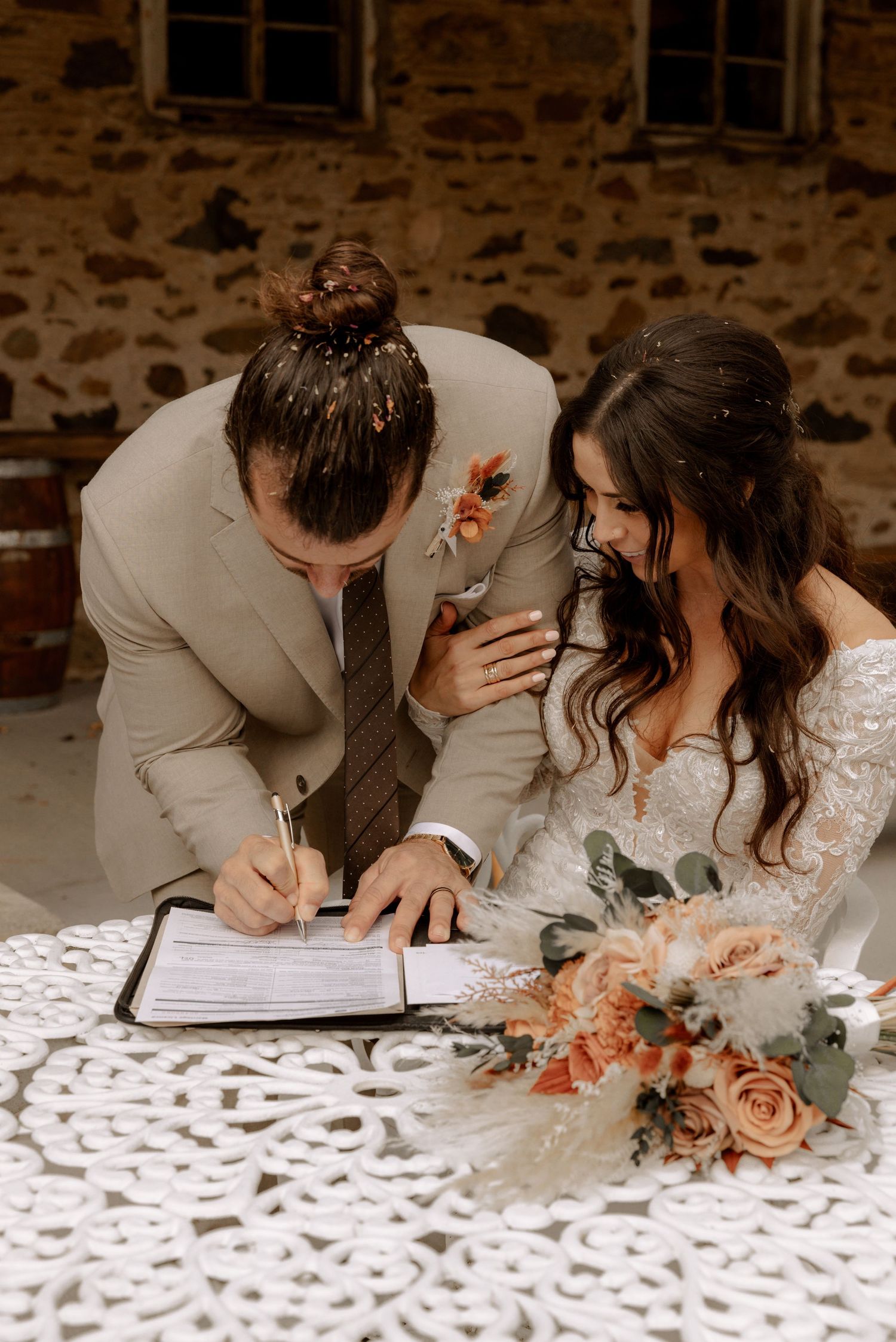 A couple signing their marriage license with a peach and coral floral bouquet on a lace tablecloth.