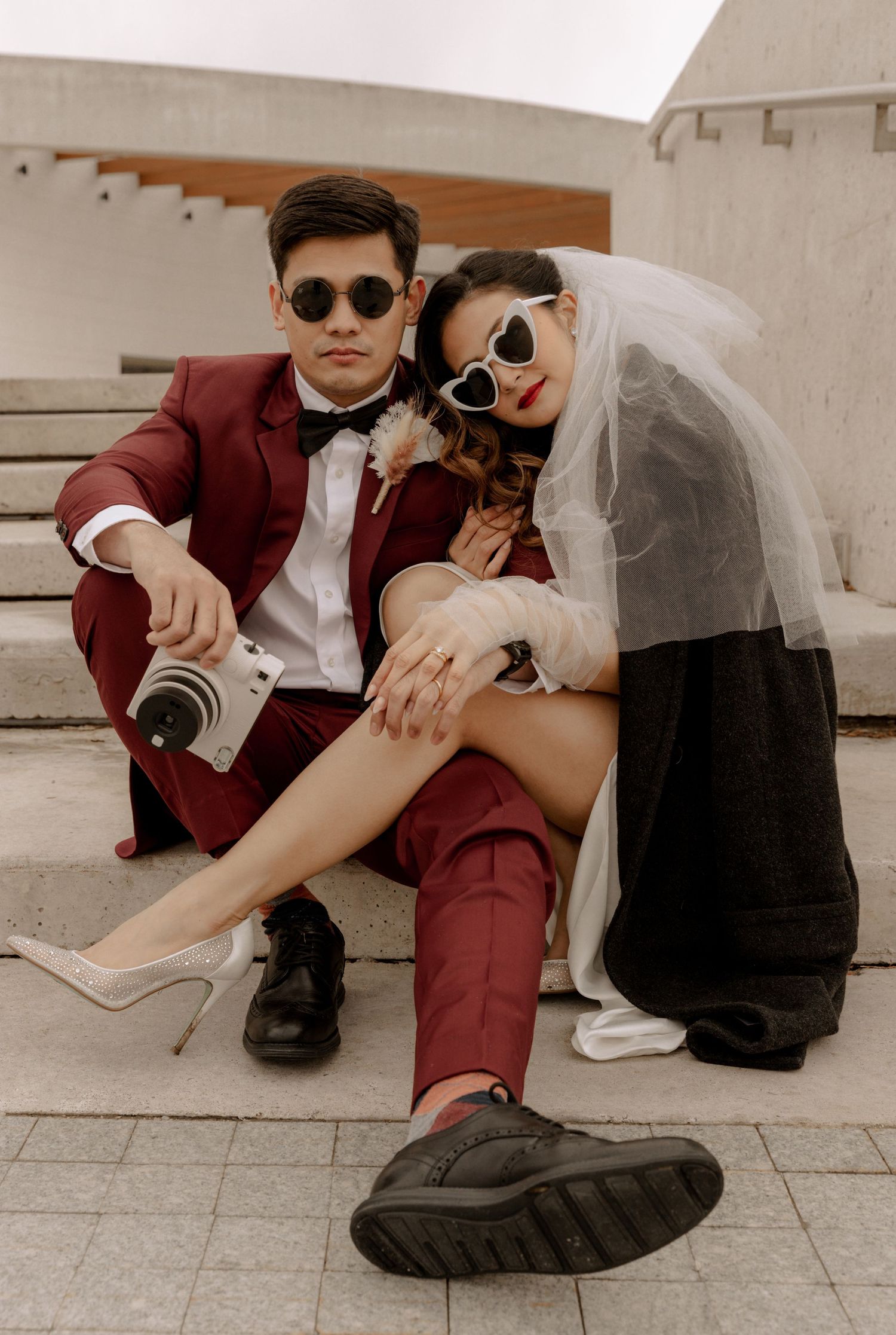 Stylish couple in burgundy suit and bridal wear pose with a camera on the ground.