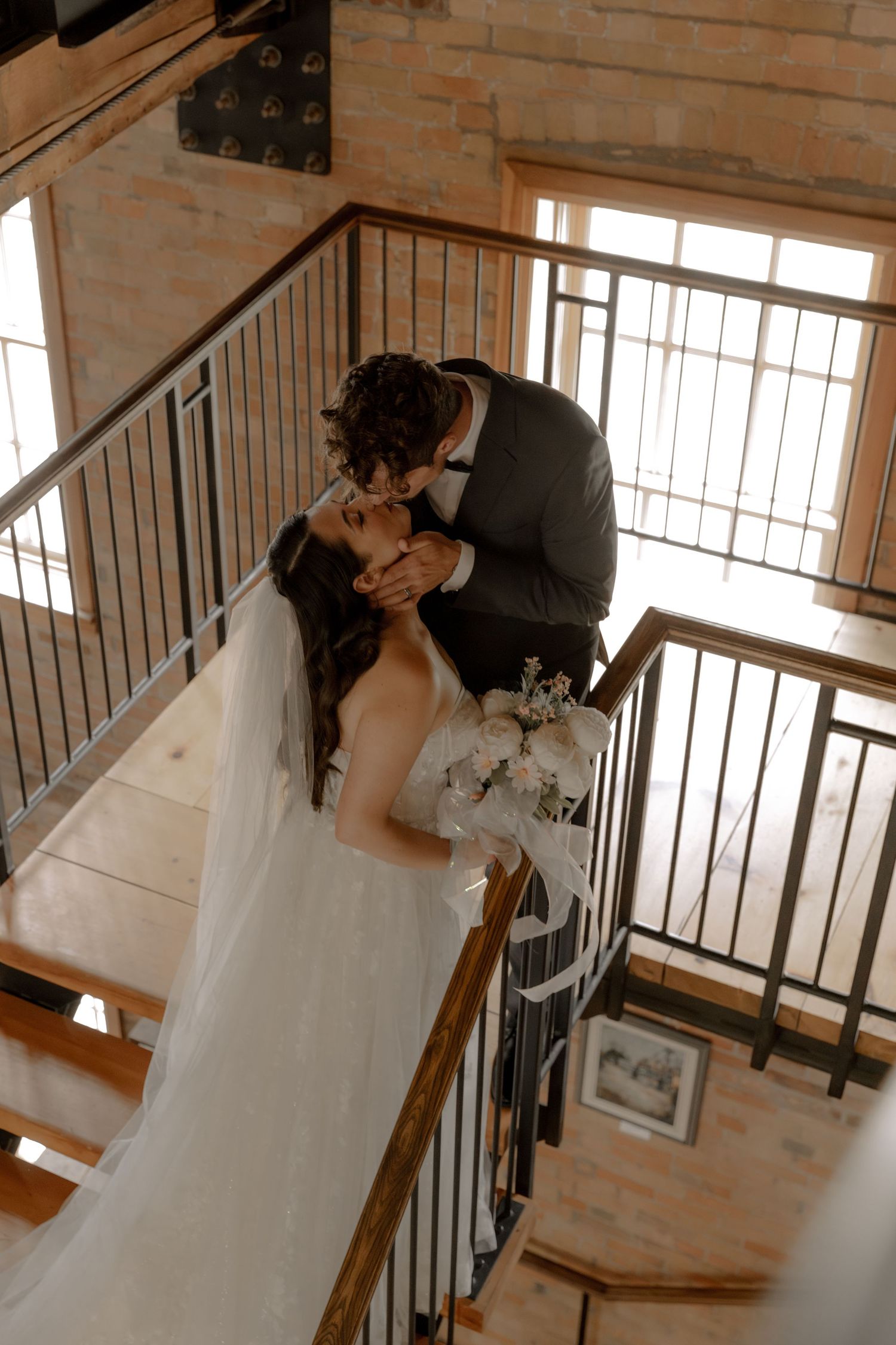 Romantic wedding moment captured on wooden staircase in rustic brick interior.