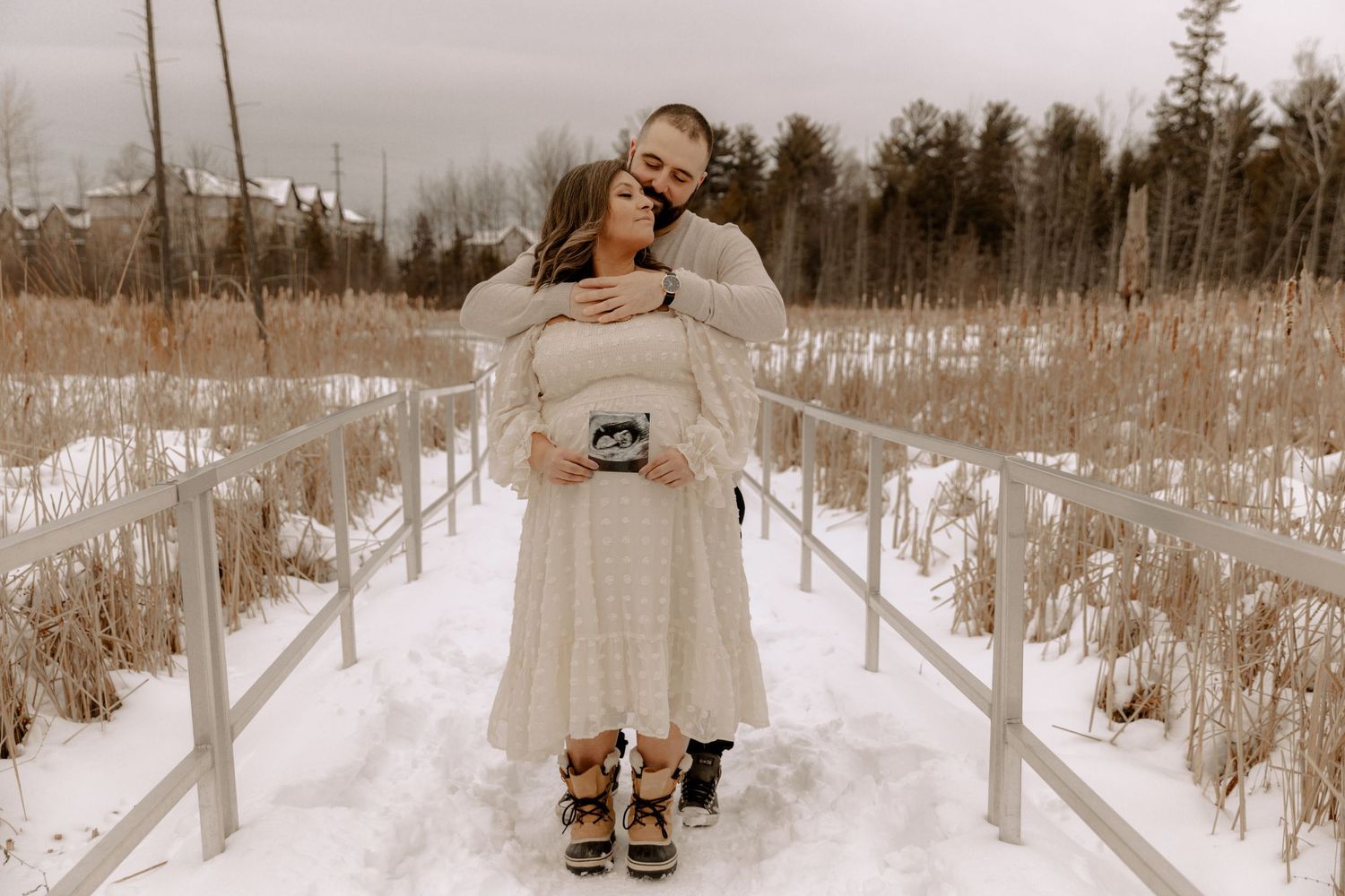 A couple embraces on a snowy walkway surrounded by winter prairie grass and bare trees in a sepia-toned photograph.