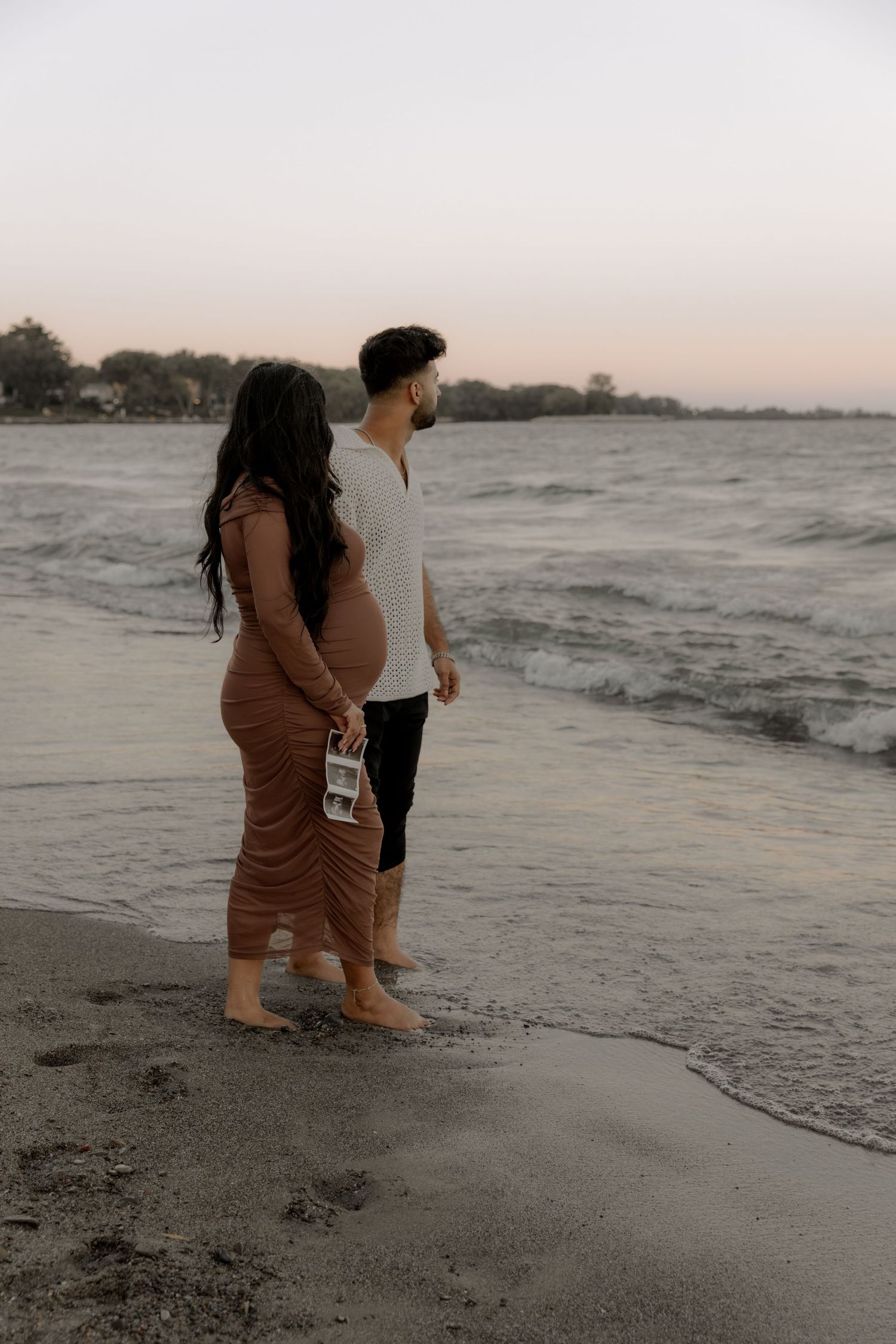 A couple stands together on the beach at dusk as waves lap at the shore while holding ultrasound photos.