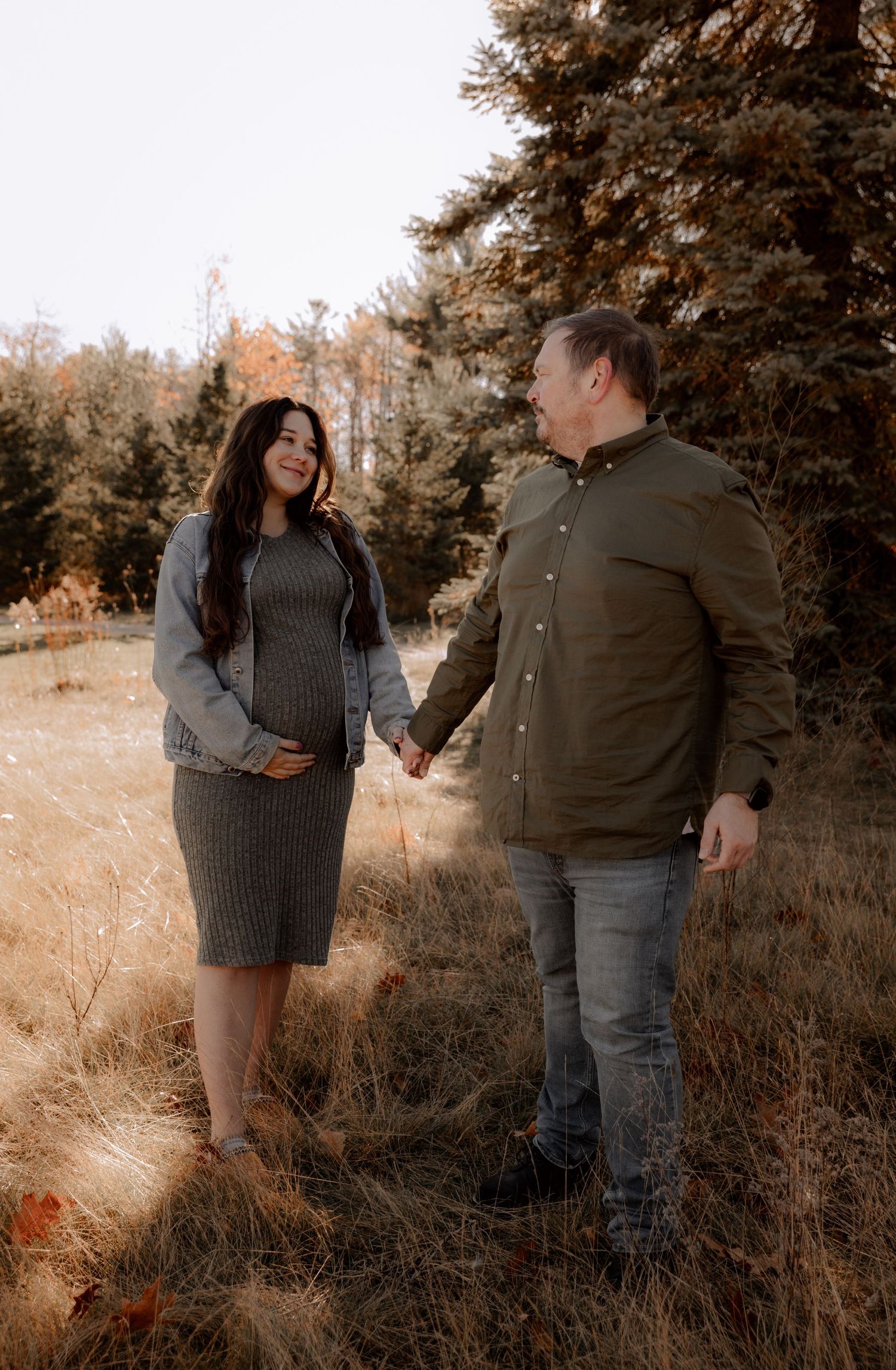 A couple holding hands in an autumn field with brown grass and fall foliage in the background on a sunny day.