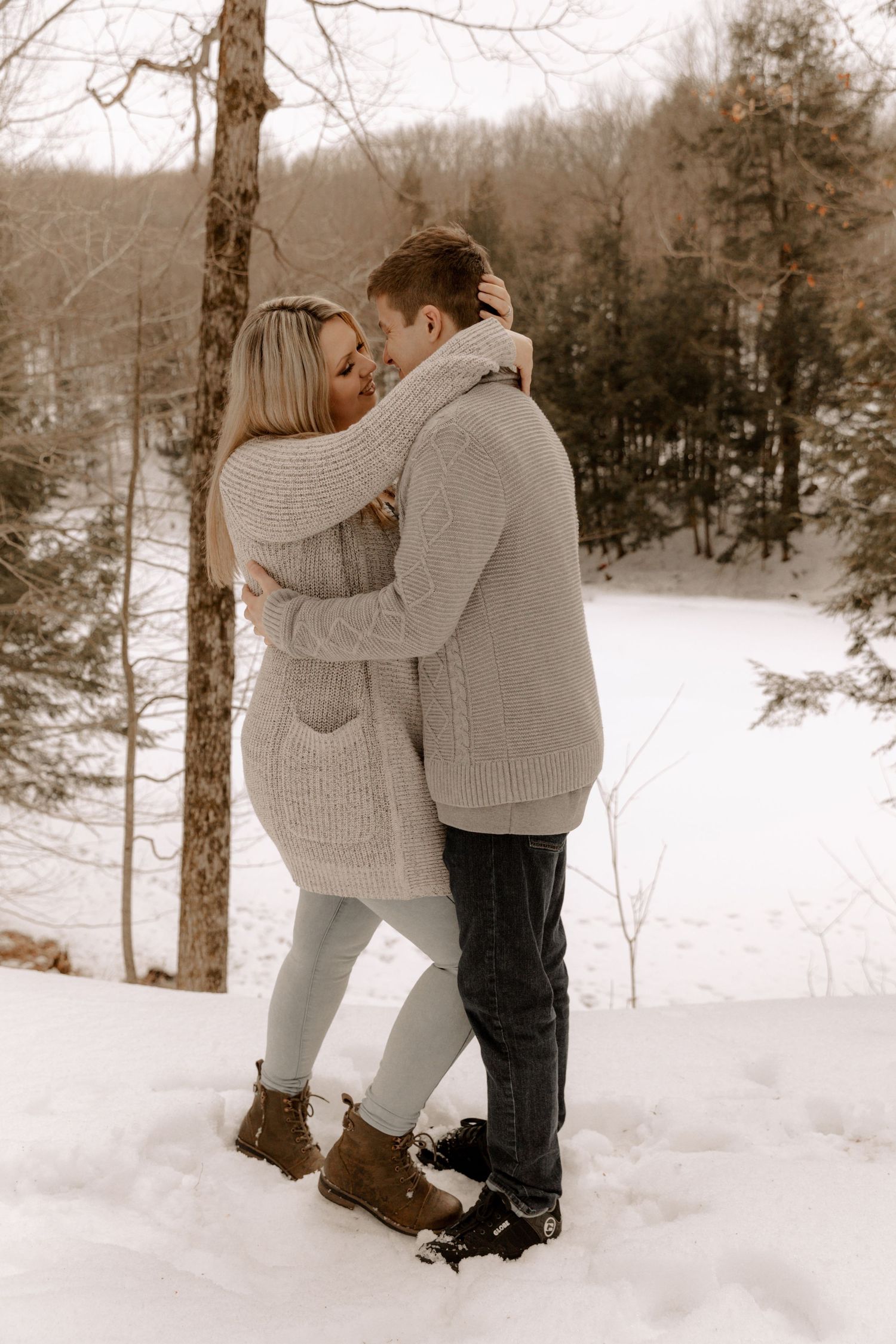 A couple in cozy sweaters embrace in a snowy winter forest setting, creating a romantic moment against the white landscape.