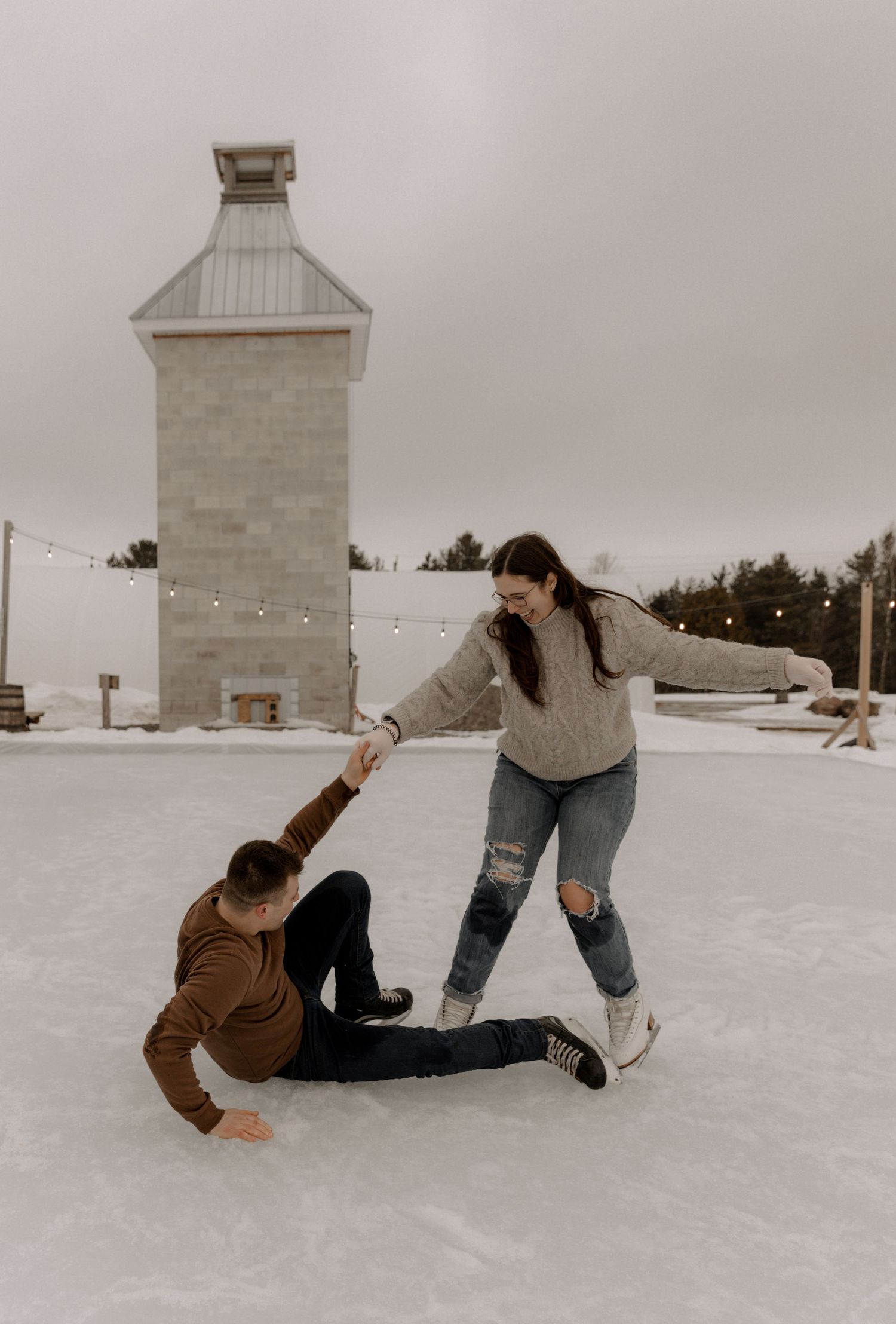 Two people ice skating outdoors at a rink with string lights and a lighthouse tower in the background.