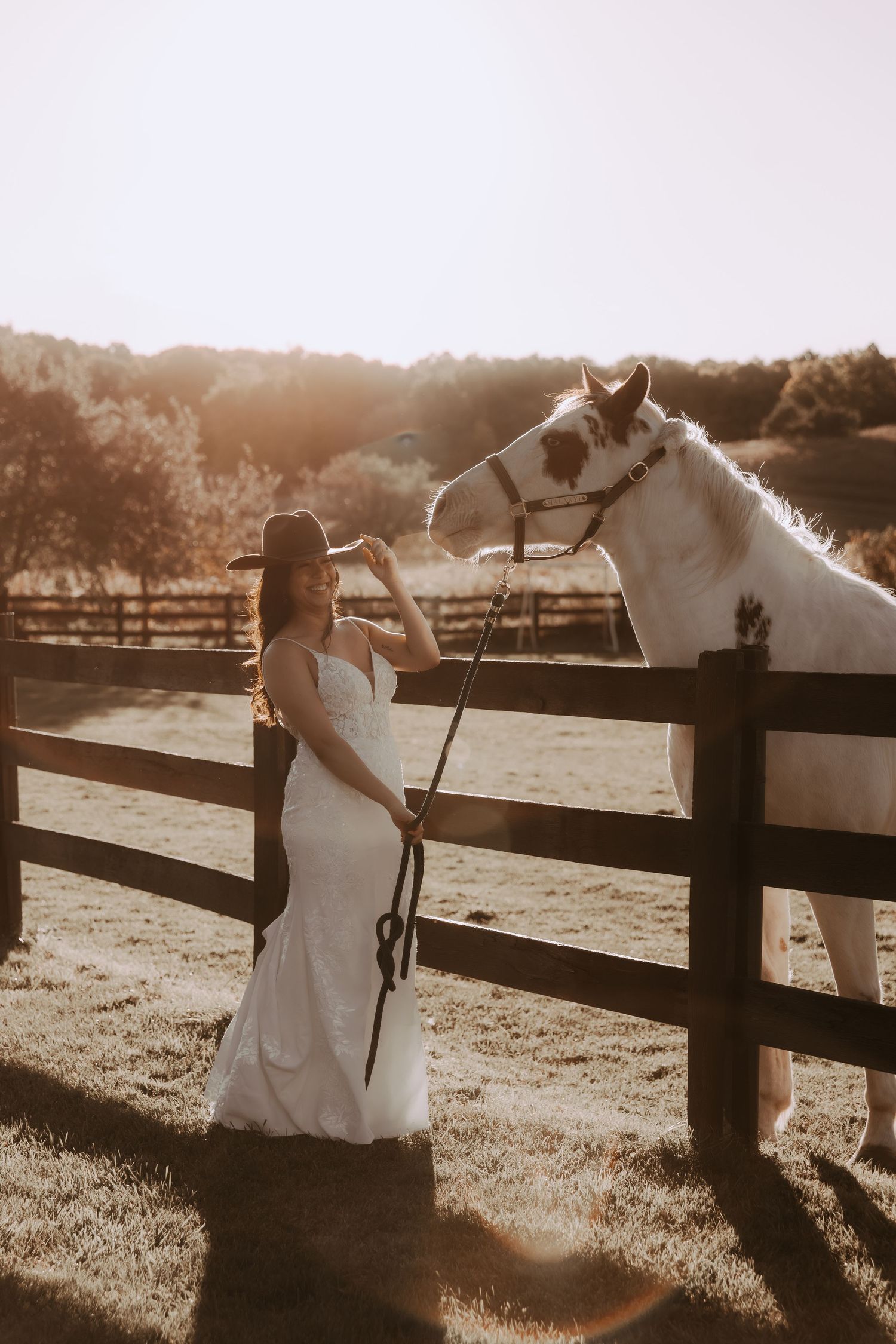 A white horse interacts with someone in a wedding gown by a wooden fence during golden hour.