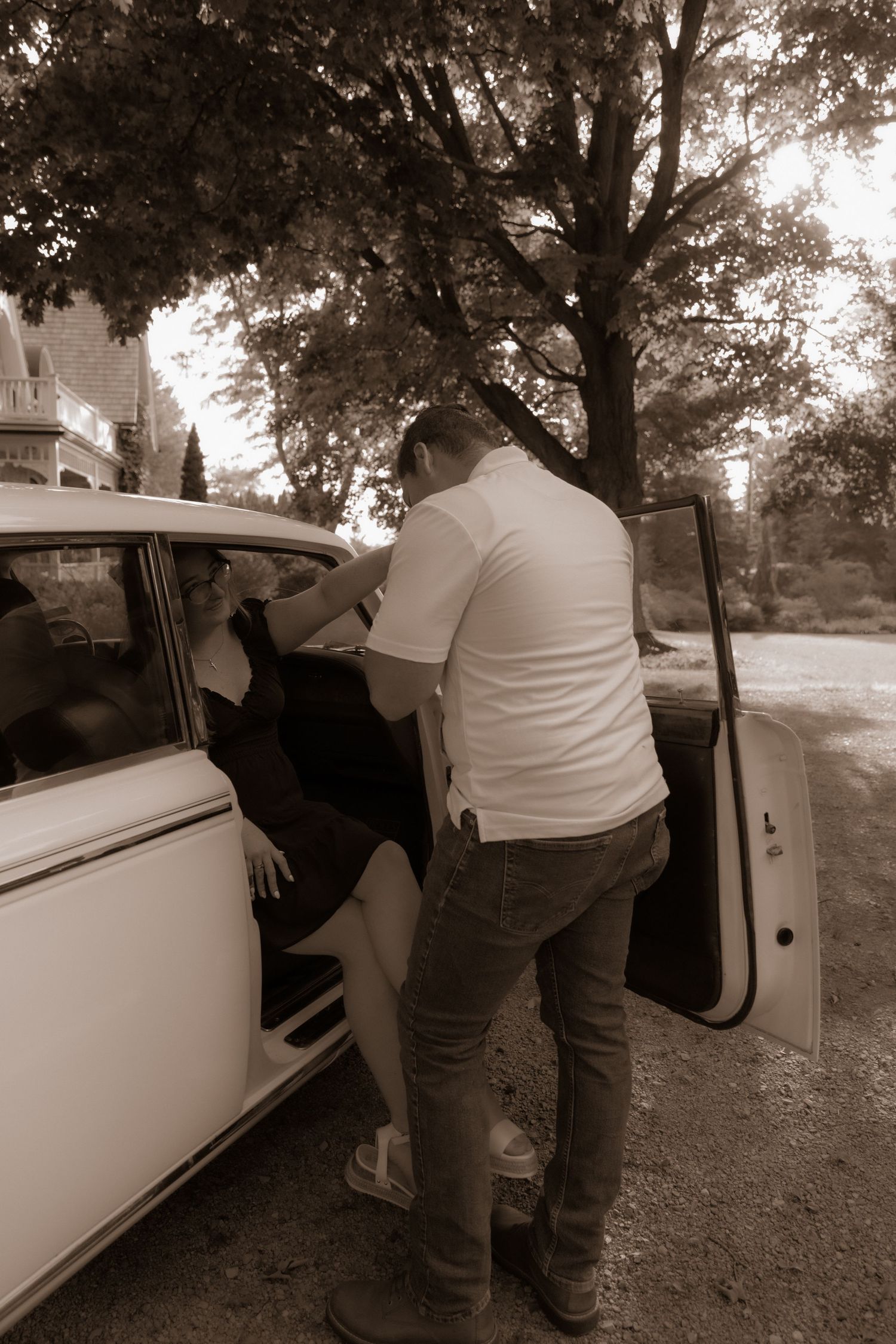 A figure in jeans and white shirt leans into vintage car door on tree-lined street in sepia-toned photograph.