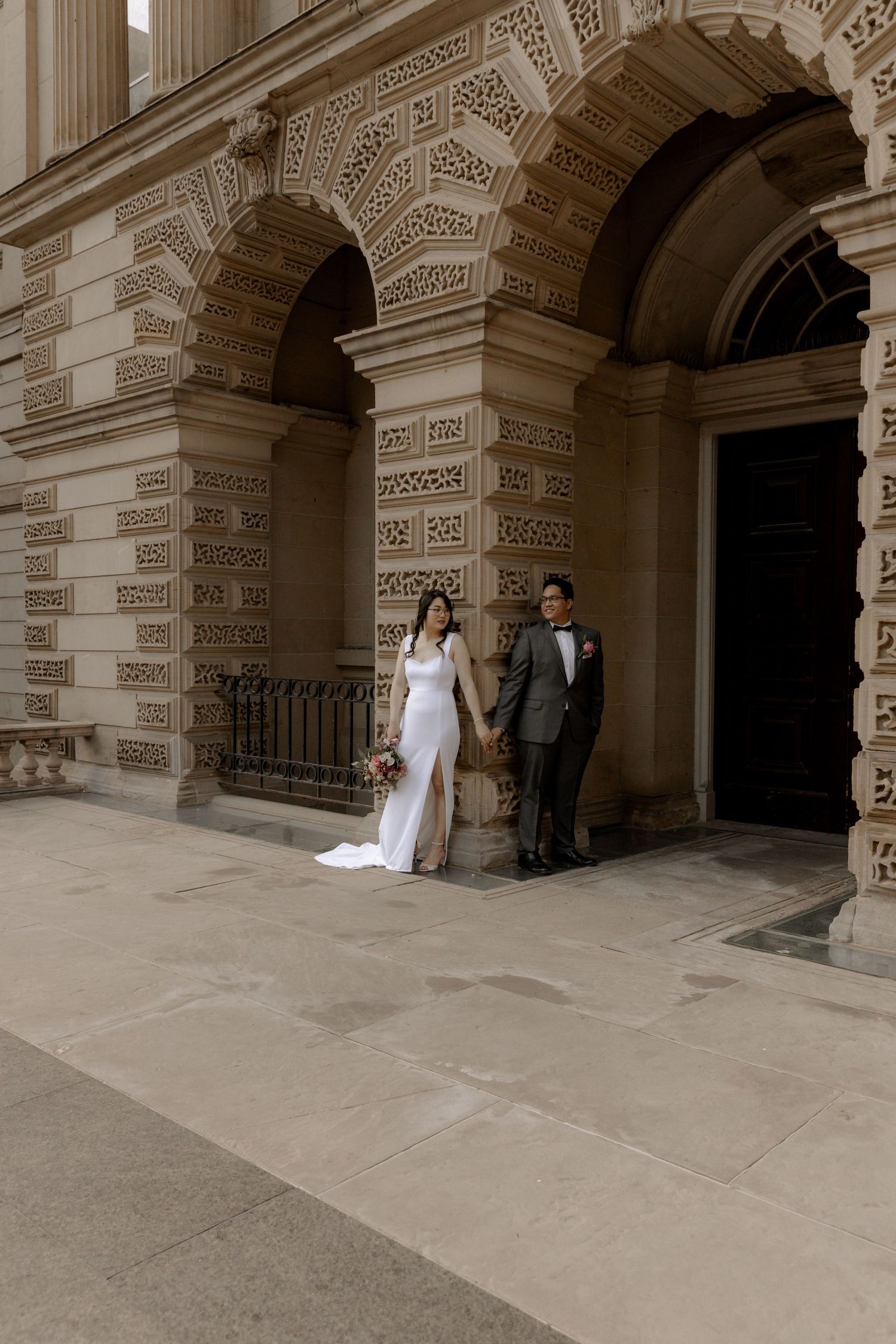 A wedding couple poses under ornate stone arches of a classic building.