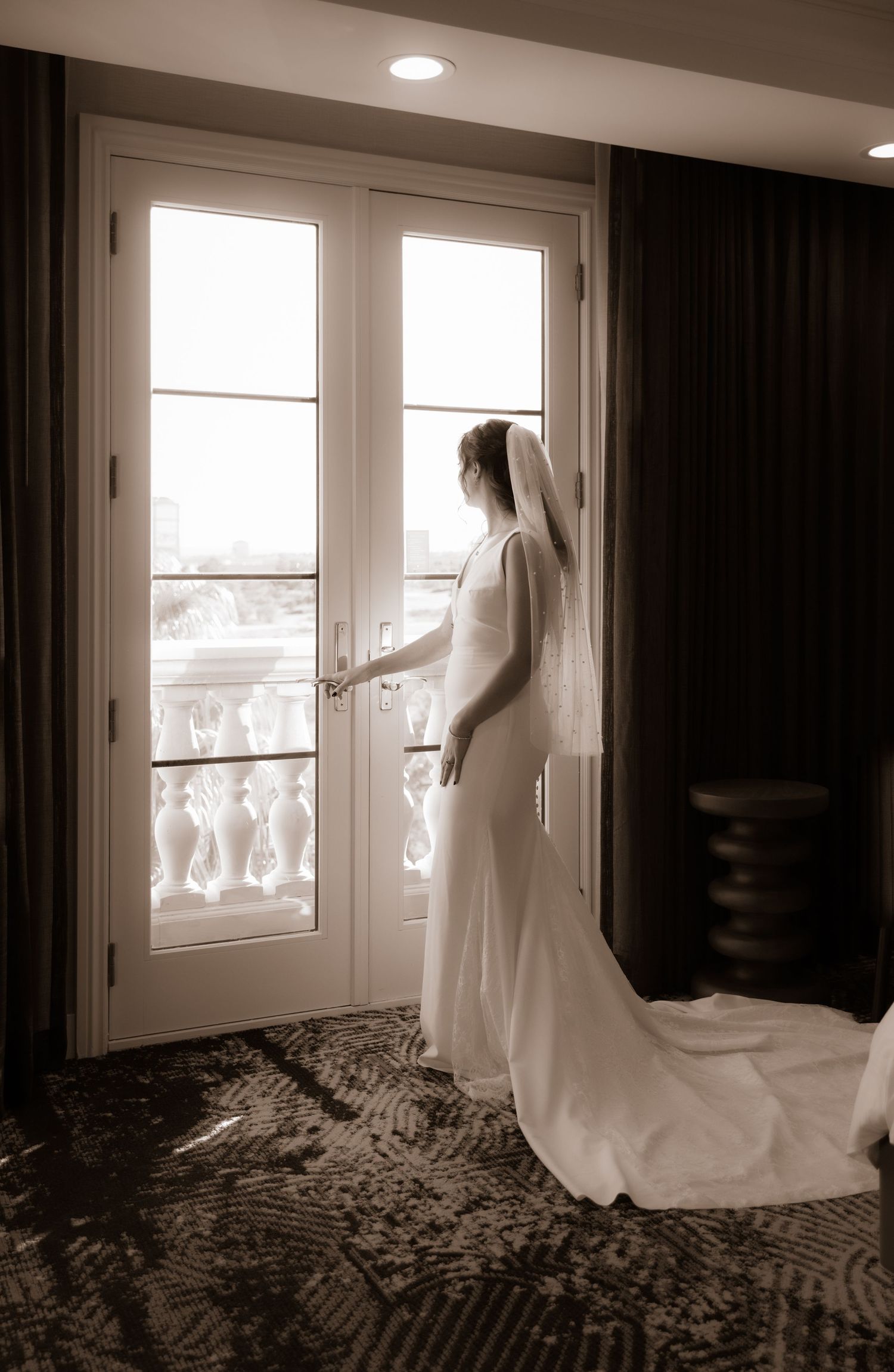 A bride in a flowing white gown stands elegantly by French doors in natural light.
