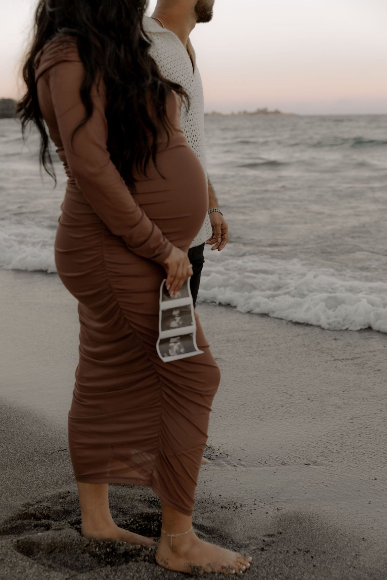 Side view of pregnant woman in brown dress holding ultrasound photos by ocean.