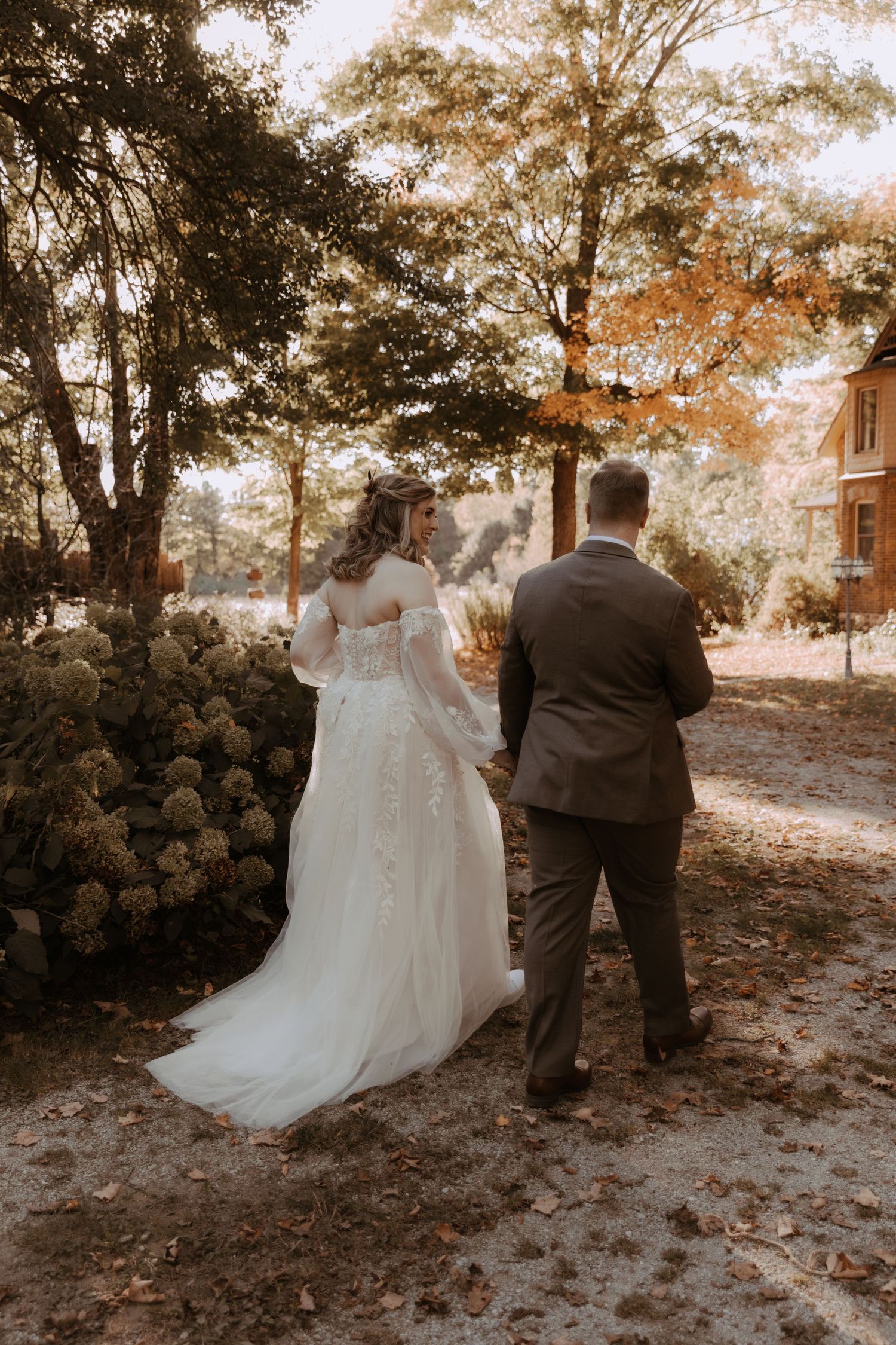 Couple in wedding attire standing outdoors in an autumn setting with sunlit trees.