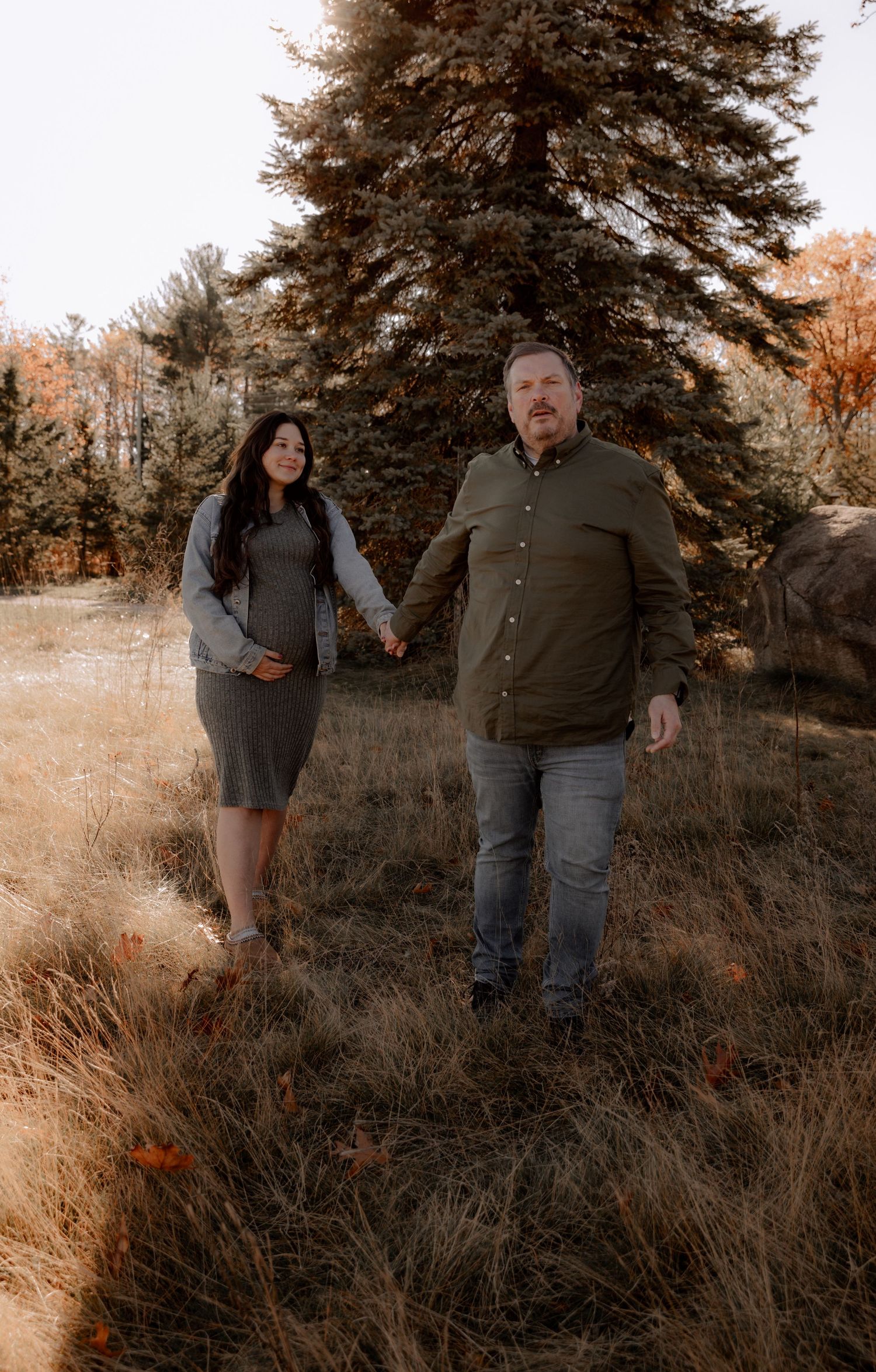 A couple stands together in an autumn field with tall grass and a large evergreen tree in the background.