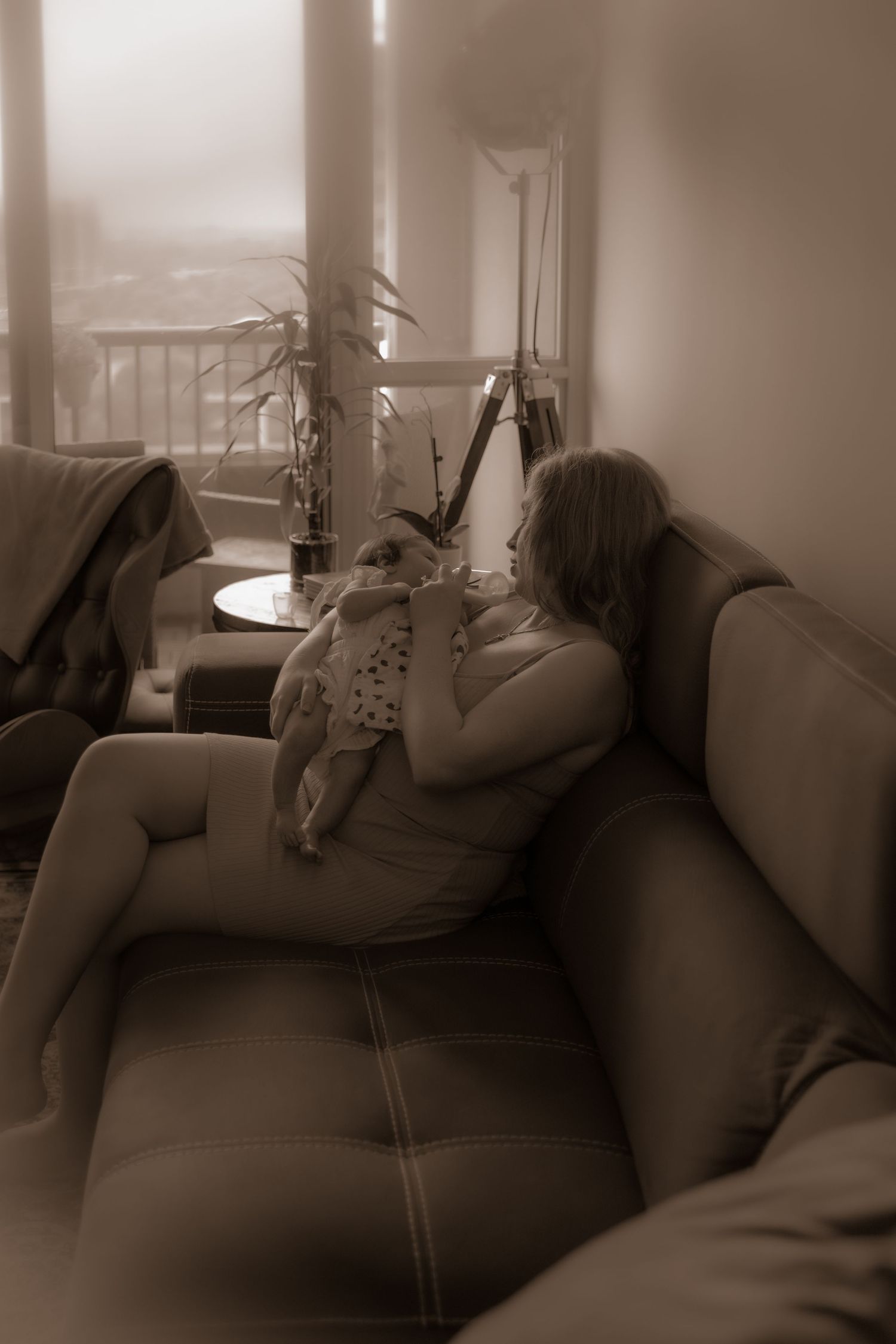 A sepia-toned image showing an intimate moment between parent and infant on a leather sofa near a window.