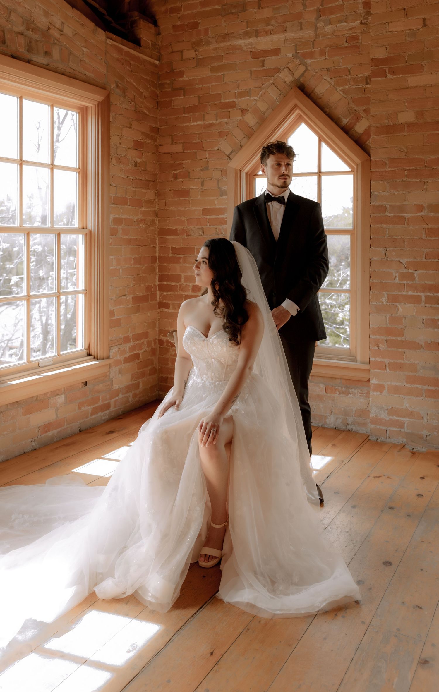 Elegant wedding couple posing in sunlit brick room with gothic windows.