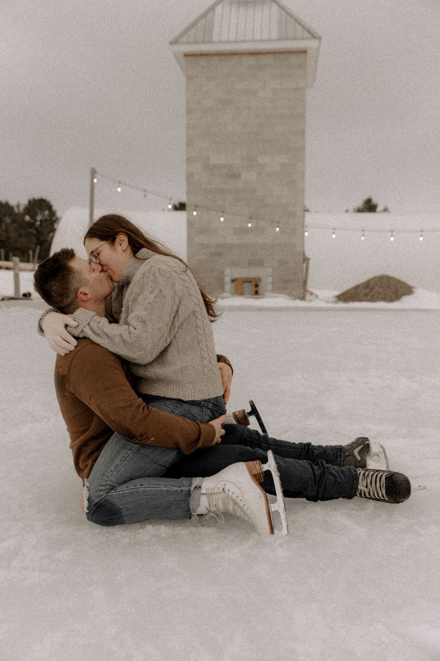 A couple sharing an intimate winter moment while sitting on snow-covered ground with string lights and building in background.
