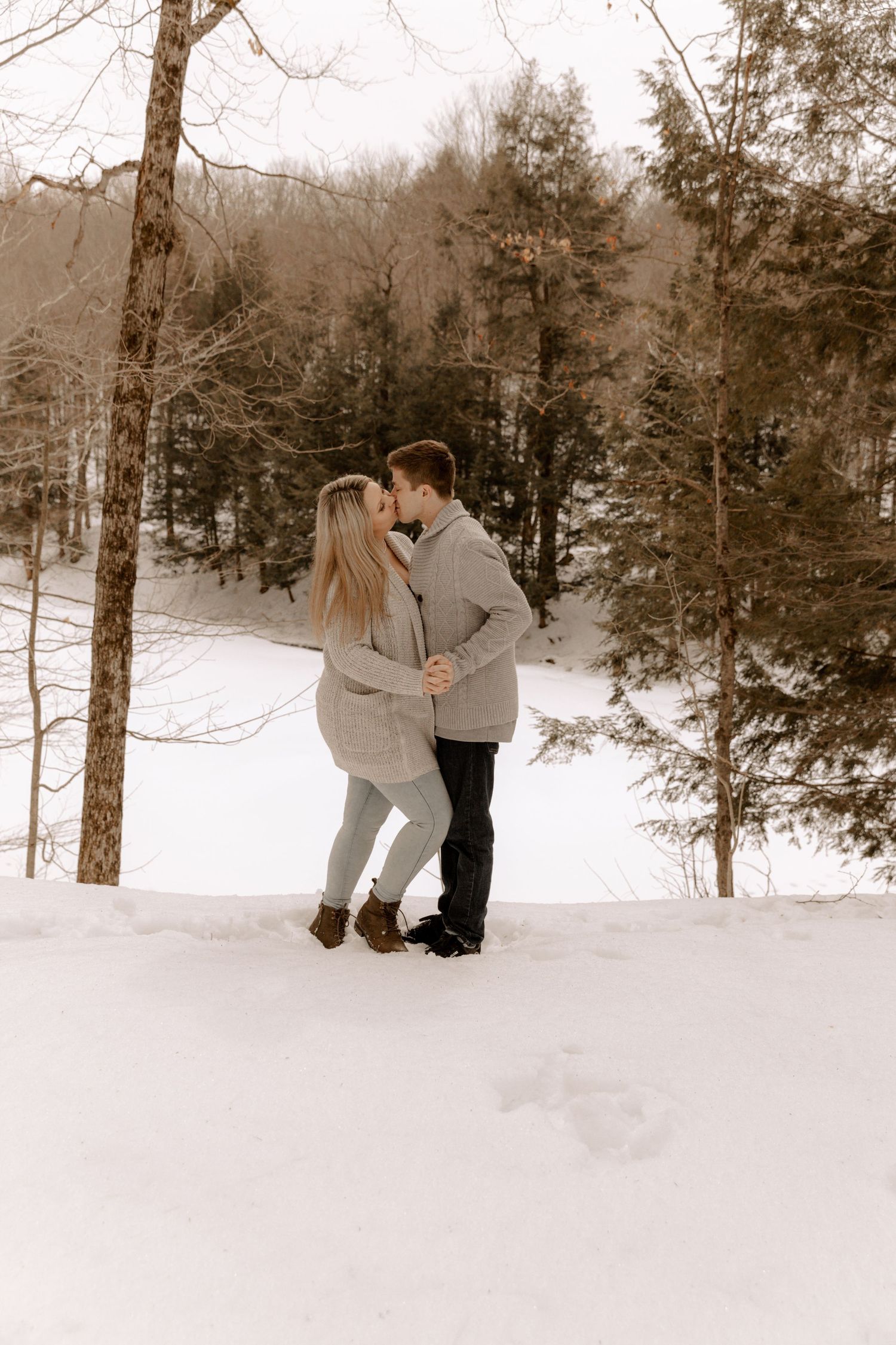 A couple embraces in a snowy winter forest scene with bare trees and evergreens in the background.