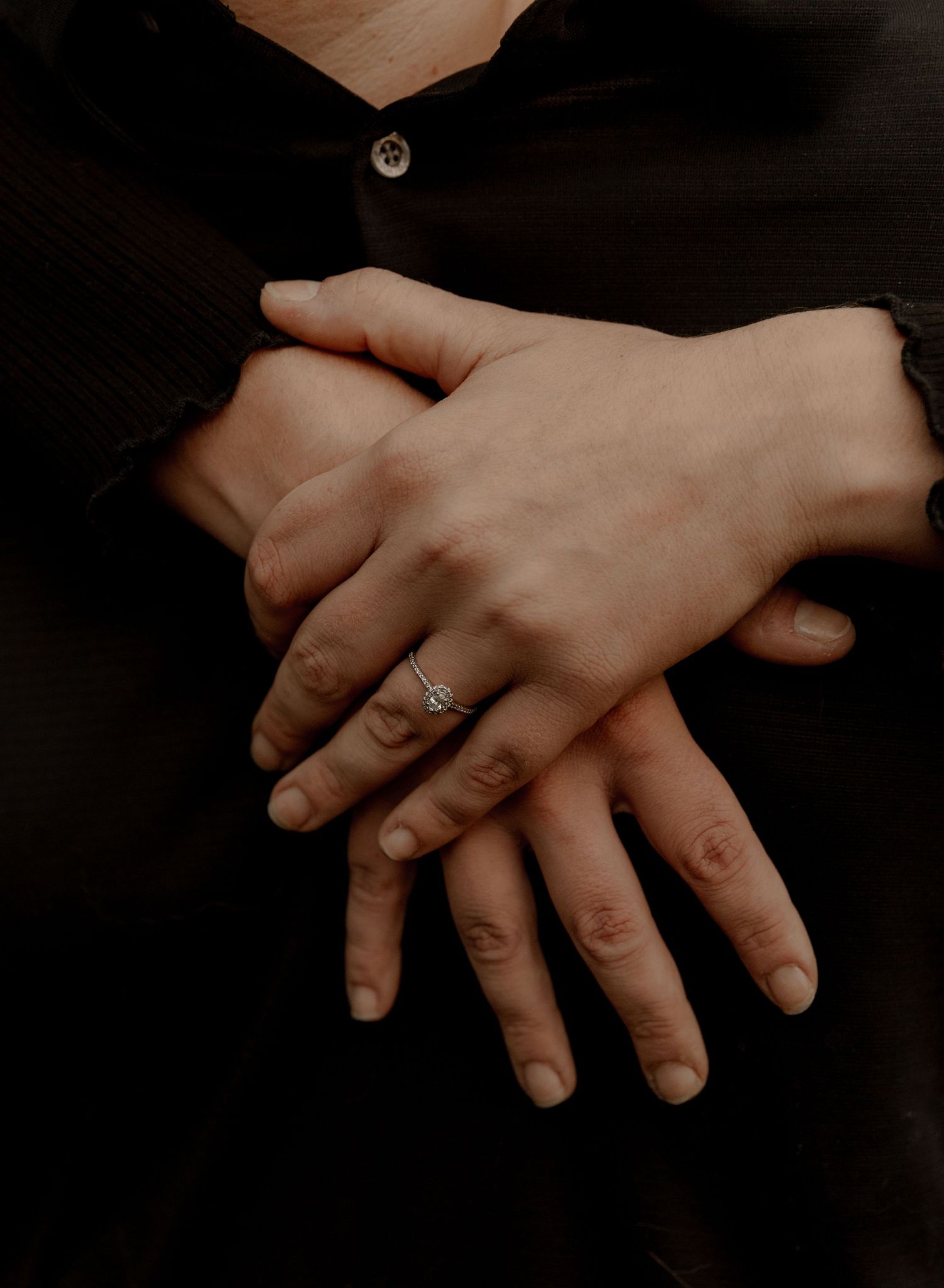 Close-up of hands with engagement ring resting on black fabric.