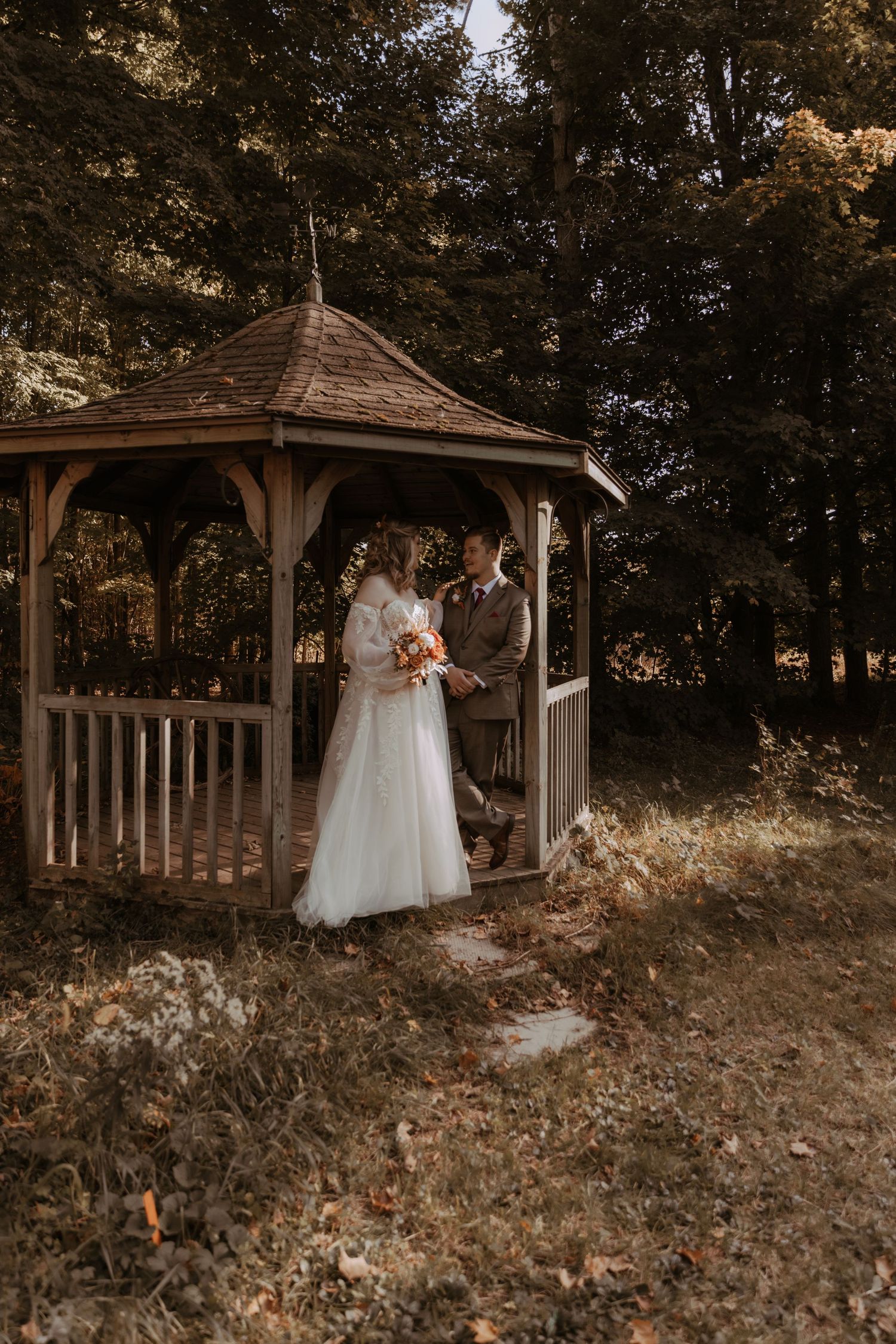 A wedding couple shares an intimate moment in a rustic wooden gazebo surrounded by autumn trees.