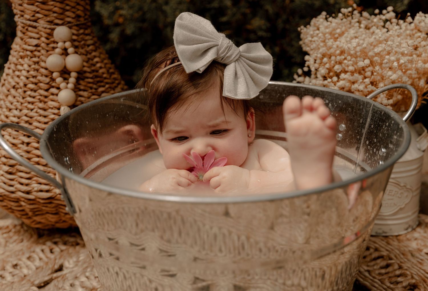 Sweet baby bathing in vintage metal tub with bow accessory and cozy beige textiles in background.