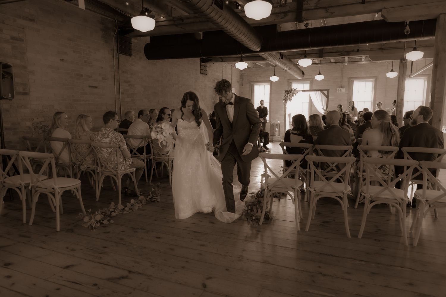 A sepia-toned wedding ceremony in a rustic venue with exposed brick walls and wooden cross-back chairs.