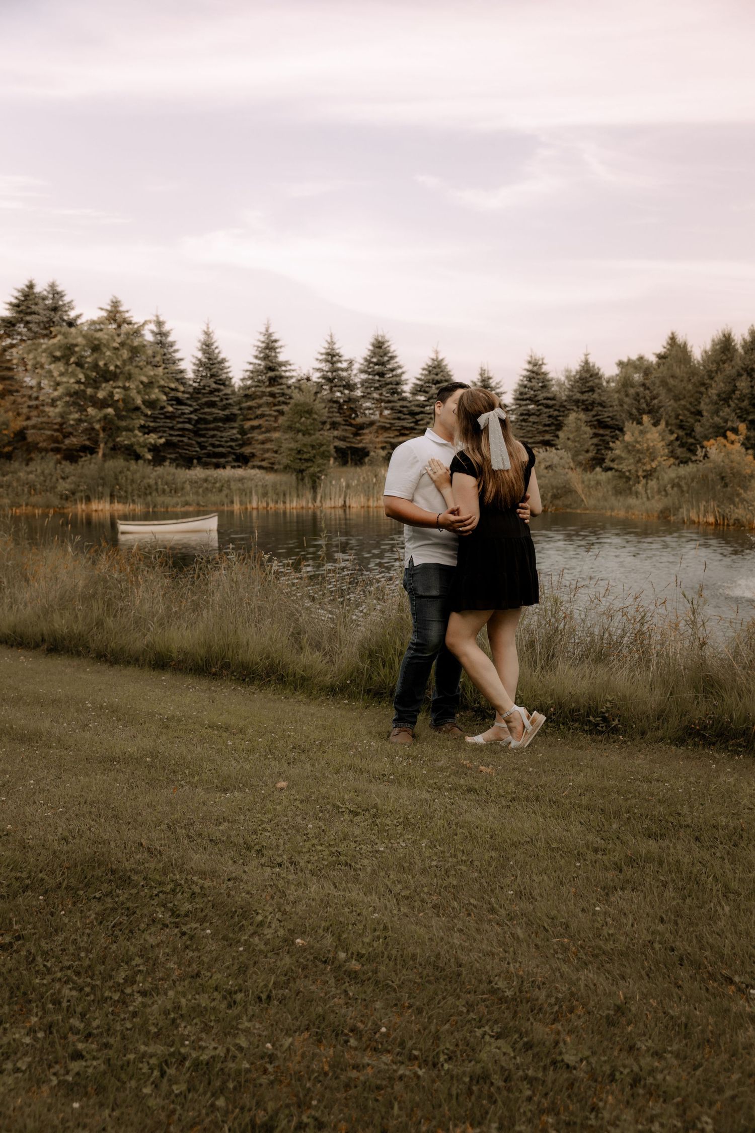 A romantic couple embraces by a serene pond with evergreen trees in the background during sunset.