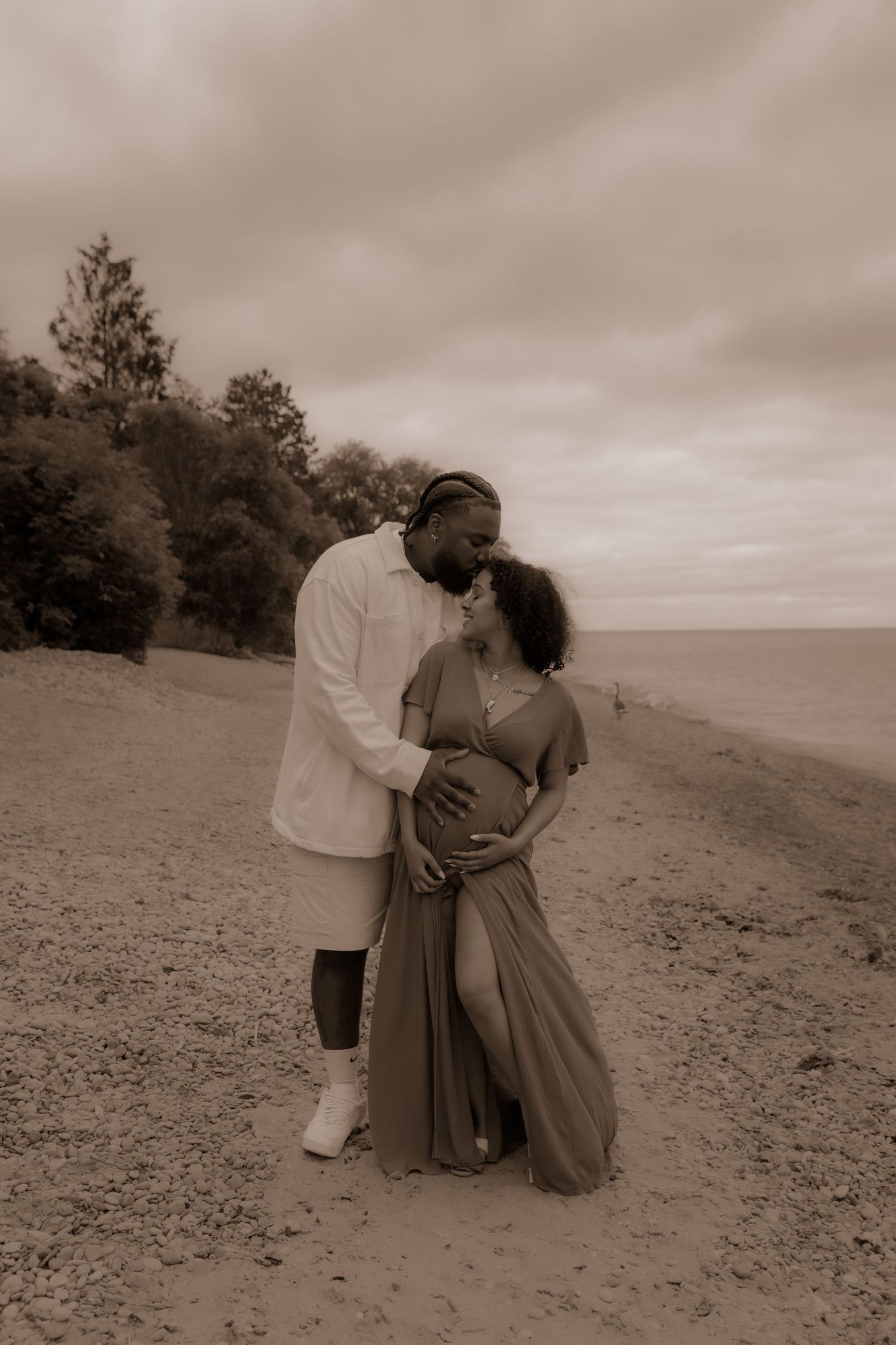 A couple embraces on a serene beach with trees in the background, captured in sepia tones.