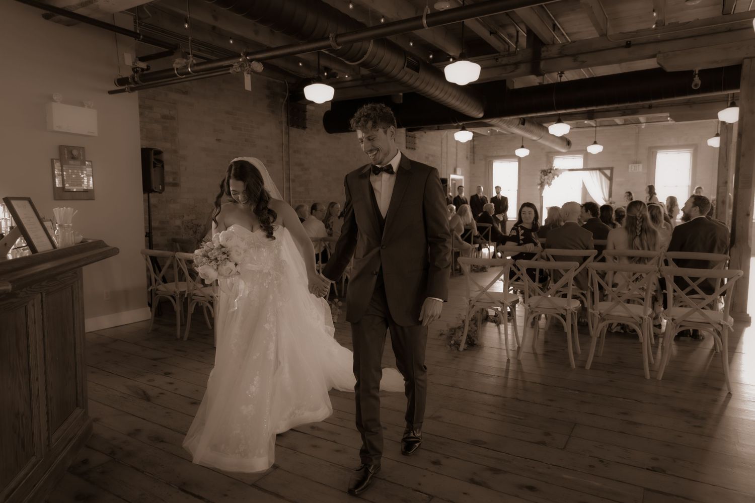 A sepia-toned photo of newlyweds walking through an indoor venue with vintage decor.
