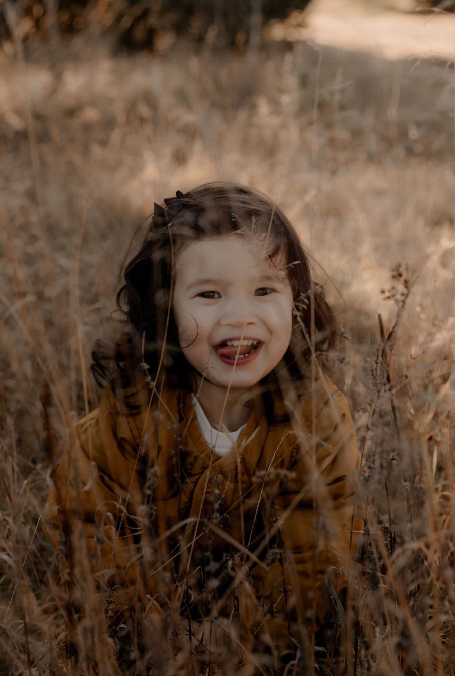 A child in a mustard sweater smiles brightly while sitting in tall autumn grass.