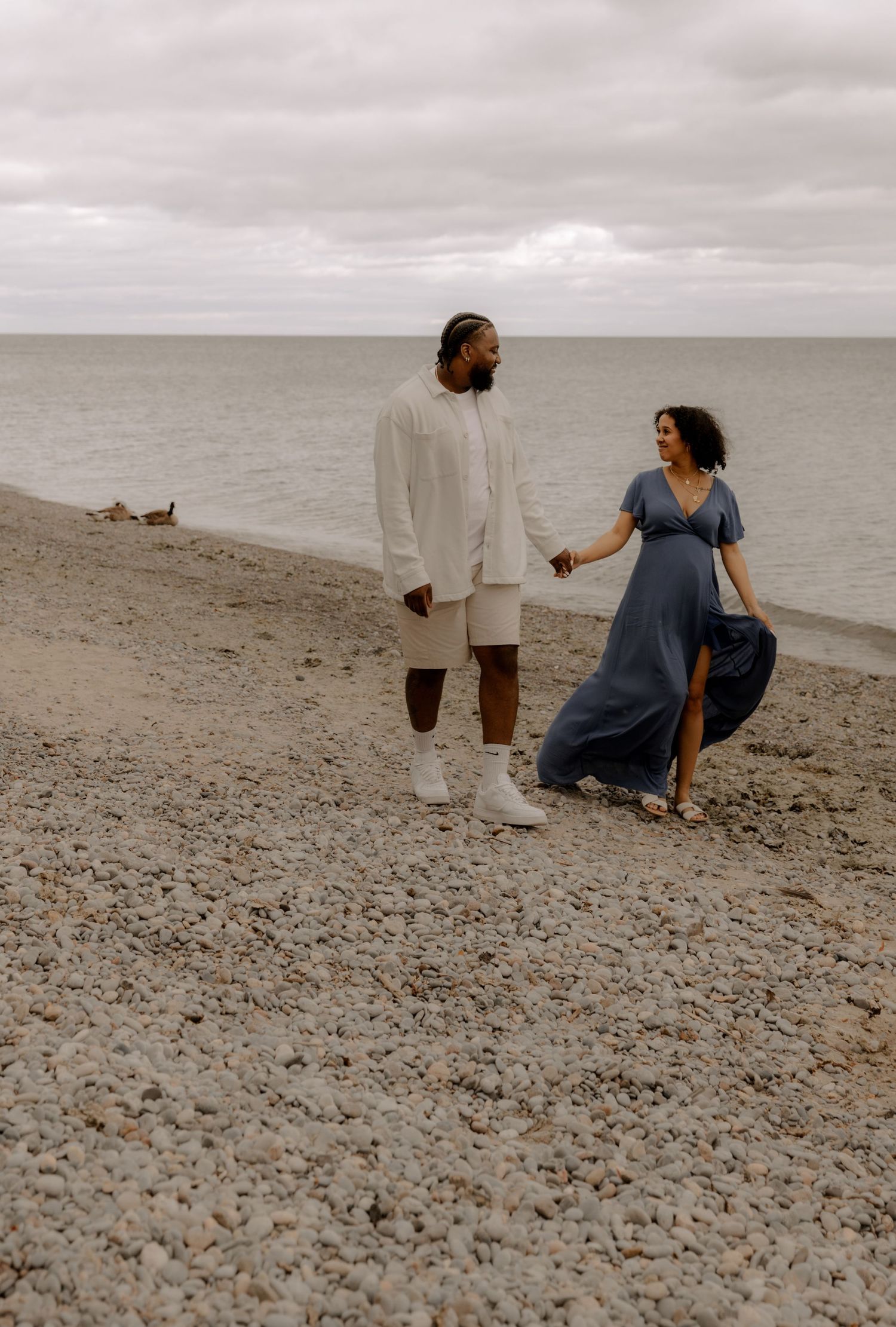 Couple walking along rocky beach shore under cloudy sky with ocean view.